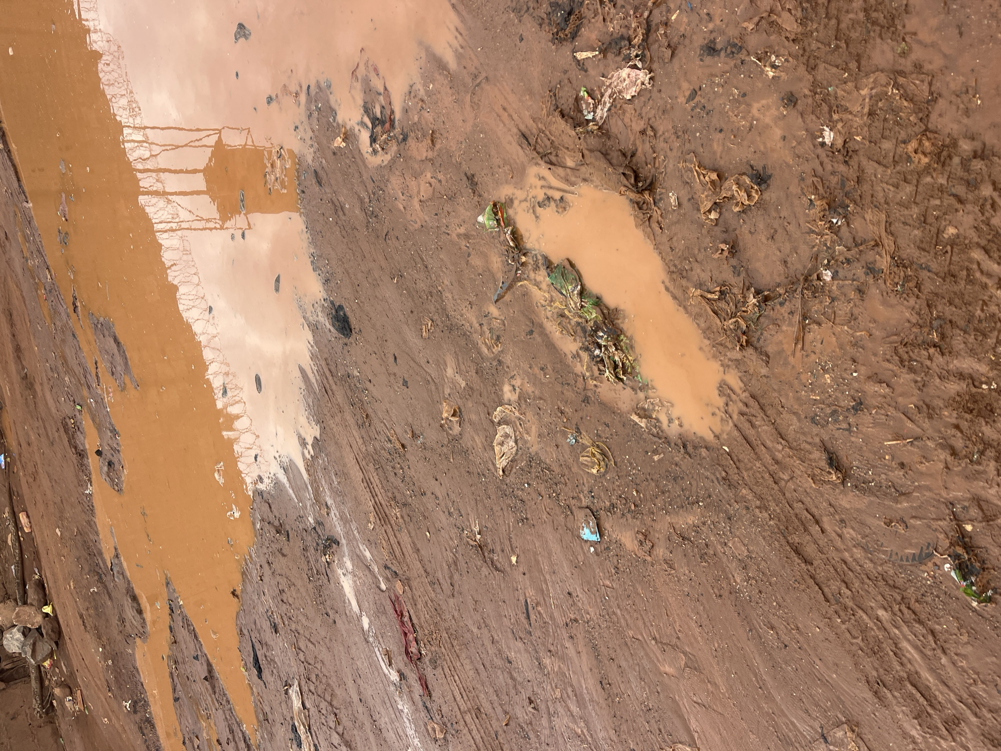 The image shows a muddy, trash-strewn area. There is standing water with a reddish-brown hue, suggesting the presence of iron oxides in the soil. The water reflects the vague outline of a structure with what appears to be a chain-link fence. Scattered debris is visible throughout the scene, including plastic bags and other discarded materials. The ground shows evidence of tire tracks. Specific details to pinpoint the exact location within Abuja, Nigeria are absent from the image. No people are visible in the image.