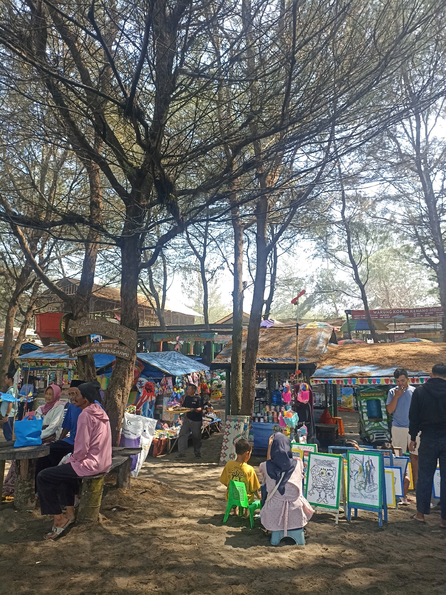 The image depicts a beachside scene in Kenoyajaya, Indonesia, characterized by numerous trees with sparse foliage, suggesting a dry or autumnal season. The ground is sandy, indicative of a beach environment. Several stalls or kiosks are set up, selling various goods, with colorful awnings providing shade.
In the foreground, a group of people are seated at a wooden table. A woman wearing a pink headscarf and a pink long-sleeved shirt is on the left, with a blue bag placed on the table. Beside her, a person in a blue shirt and a person in a pink hooded jacket are also seated.
Further back, a variety of stalls are visible. One stall has a sign that reads "WARUNG KOLAM RENANG HOKI," indicating it is a food and drink establishment, possibly near a swimming pool. Another stall displays items for sale, including toys and accessories.
In the middle ground, a young boy in a yellow t-shirt is seated on a green plastic chair, facing a woman in a light purple headscarf and a patterned dress. The woman is seated on a blue stool, looking at what appears to be a drawing board. Several other drawing boards are displayed in front of them, some with cartoon characters like SpongeBob SquarePants and a bulldozer depicted. Other children and adults are also present in the vicinity of the stalls.
In the background, more trees and structures are visible, including what appear to be buildings and more market stalls. The sky is bright and hazy, suggesting a sunny day. The overall atmosphere appears to be a lively marketplace or recreational area on a beach.