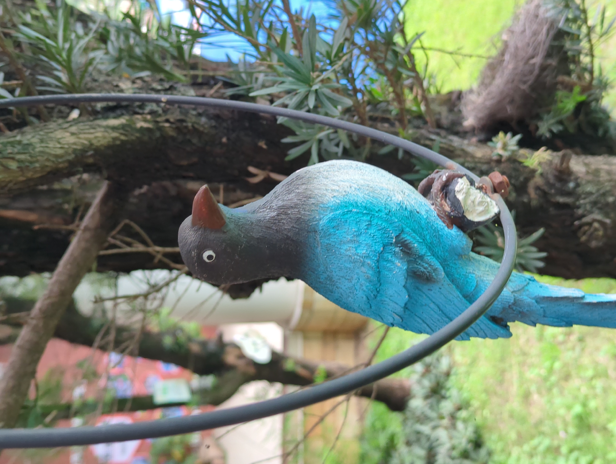 A low-angle, close-up shot depicts a weathered, turquoise-blue bird ornament suspended from a gray metal hoop. The bird's head is black with a small, red horn-like projection above its eye. The setting includes tree branches with dense, green foliage. The background features indistinct greenery, suggesting an outdoor or garden setting. The exact location in Shunde District, China, is not identifiable from the image.