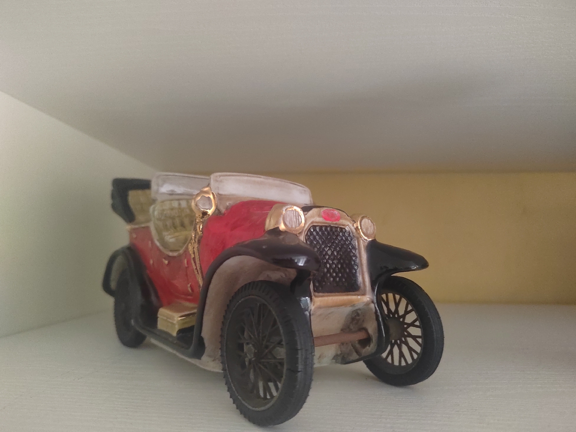 A ceramic figurine of a vintage red and black convertible car is displayed on a white shelf against a blurred light beige background. The car has gold accents around the headlights, grille, and detailing. Its wheels are black with spokes. The figurine appears to be a decorative item. The environment suggests an indoor setting, possibly a home or a shop, rather than a public space in Porto Recanati, Italy.