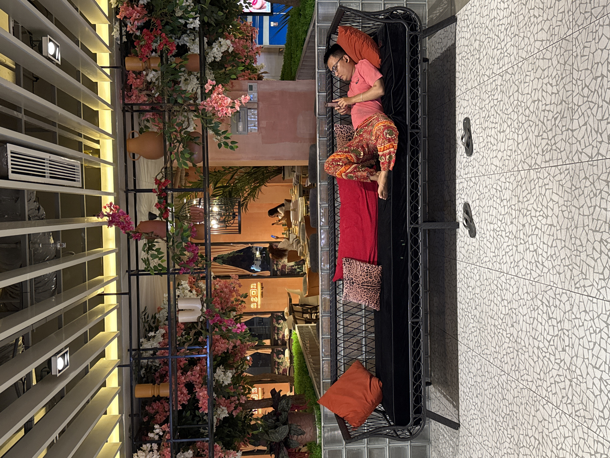 In Nha Trang Ward, Vietnam, a man is lying on a black woven couch, holding a mobile phone. He is wearing a pink t-shirt and colorful patterned pants. The couch is adorned with three pillows: one orange, one with a leopard print, and one red. In the background, a restaurant setting is visible, featuring tables, chairs, and a wall decorated with flowers. The overall environment appears to be an indoor public space with a modern, decorative design.