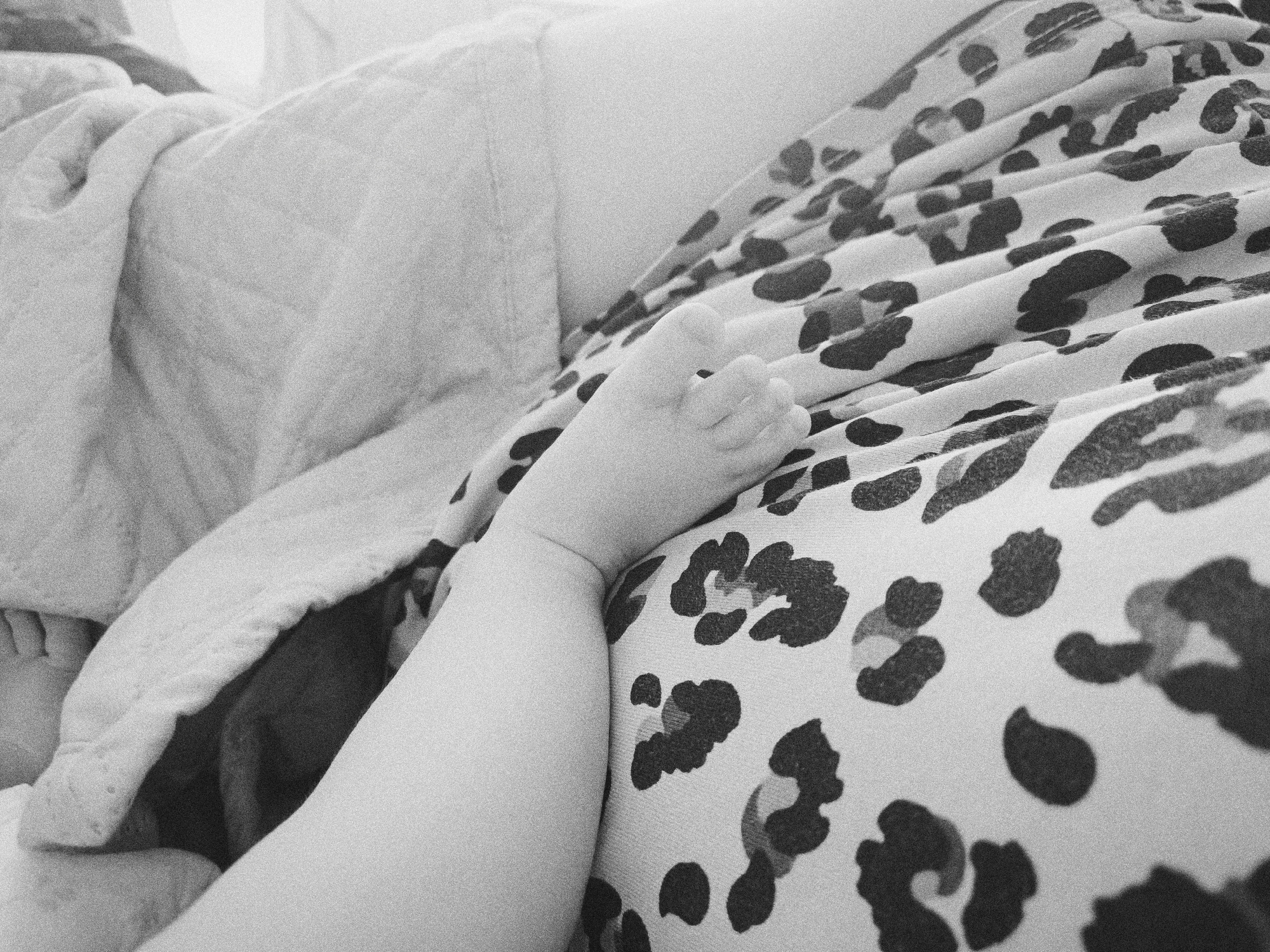 A black and white photograph depicts a close-up of a baby's foot resting on a patterned surface, likely fabric with a leopard print design. The foot is small, with discernible toes. Adjacent to the foot and fabric is a textured blanket or quilt. The background is blurred and out of focus. There is no indication of a location in Tobolsk, Russia. The focus is on the baby's foot and the adjacent textures and patterns.