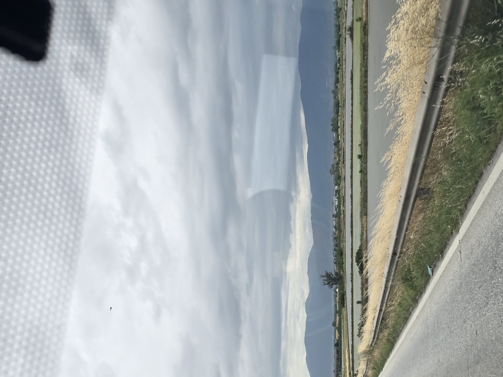 The image depicts a landscape scene with a road in the foreground, leading towards a body of water and mountains in the distance. The road is paved and has a white line marking its edge. A metal guardrail runs alongside the road, separating it from a grassy area with tall, dry grass. Beyond the grassy area is a large, flat expanse of water, possibly a flooded field or a lake, with several rectangular sections. In the background, there are low-lying hills or mountains under a cloudy sky. A single tree is visible in the middle ground, to the left of the center. The sky is overcast with dark clouds. The location is identified as Skutare, Bulgaria. There are no people or animals visible in the image. The overall impression is a rural, open landscape.