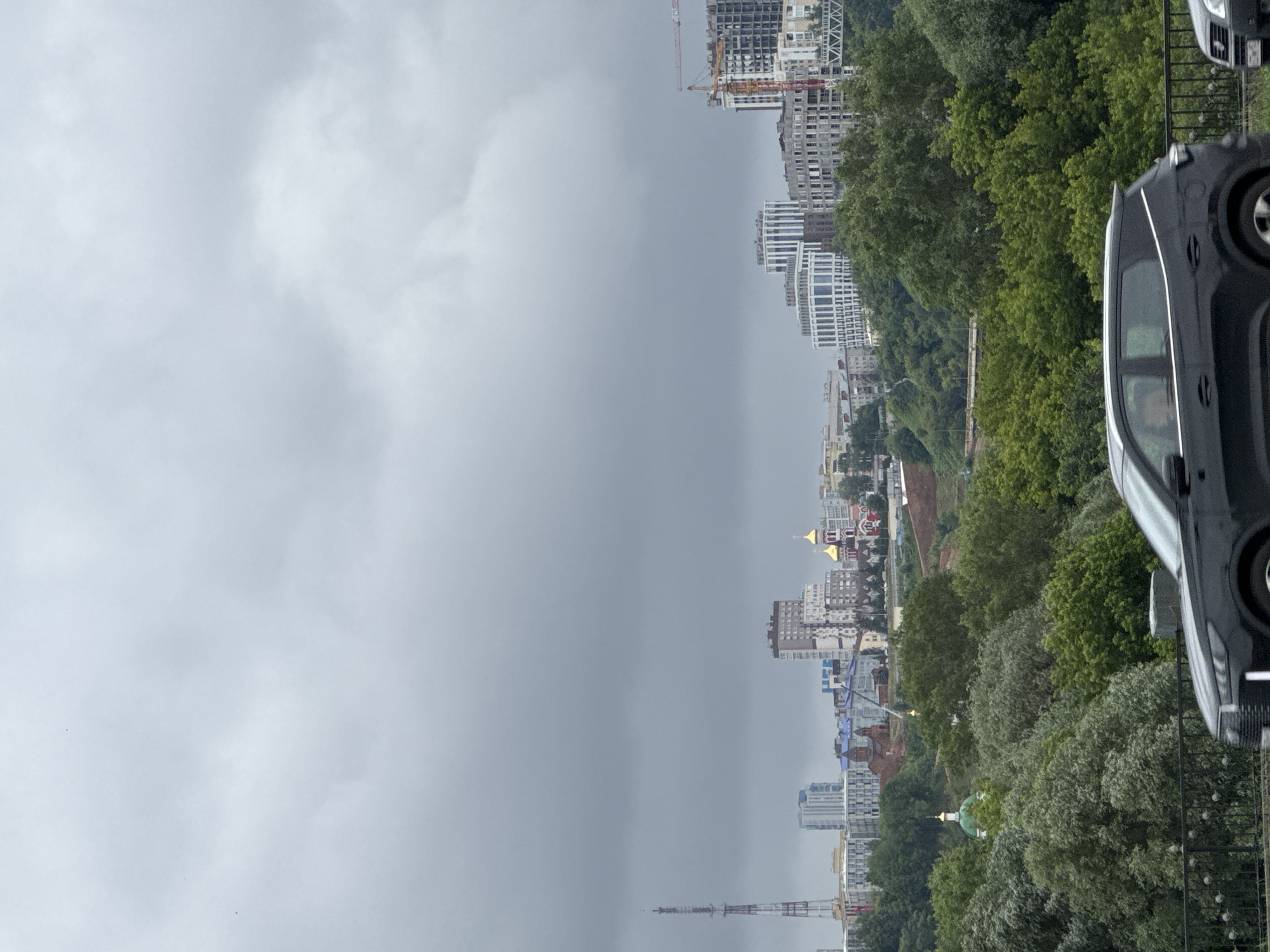 A wide shot of a city skyline on a cloudy day in Nizhny Novgorod, Russia, with a foreground of lush green trees and a dark-colored SUV driving past. The skyline features a variety of buildings, including modern high-rises, older structures, a prominent radio tower on the left, and a golden-domed church in the center. Construction cranes are visible on the right side of the skyline, indicating ongoing development. The sky is overcast with heavy gray clouds, suggesting the possibility of rain. The trees in the foreground are dense and varied in shades of green, creating a natural buffer between the viewer and the urban landscape. The SUV is the only vehicle clearly visible, with its front section in focus and its rear fading out of frame.