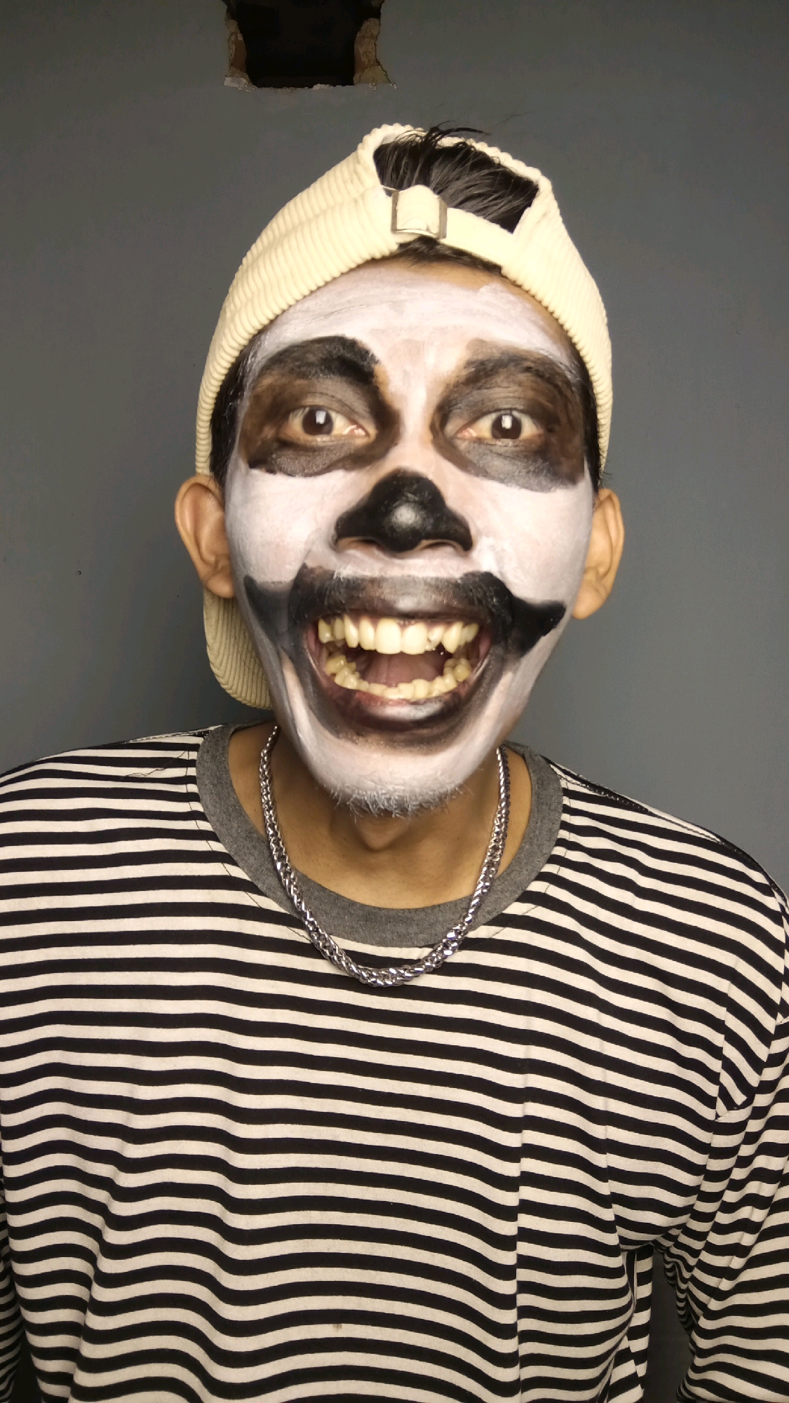 A medium shot captures a person from the chest up, their face painted in a skeletal design with white makeup covering most of their face, black around the eyes and nose, and dark lines around the mouth to simulate a grimace. The person is wearing a light-colored baseball cap and a black and white horizontally striped long-sleeved shirt with a grey collar. A silver chain necklace is visible. The background is a plain, dark grey wall with a noticeable hole near the top center. The image does not provide any specific information about the location in Kebumen, Indonesia.