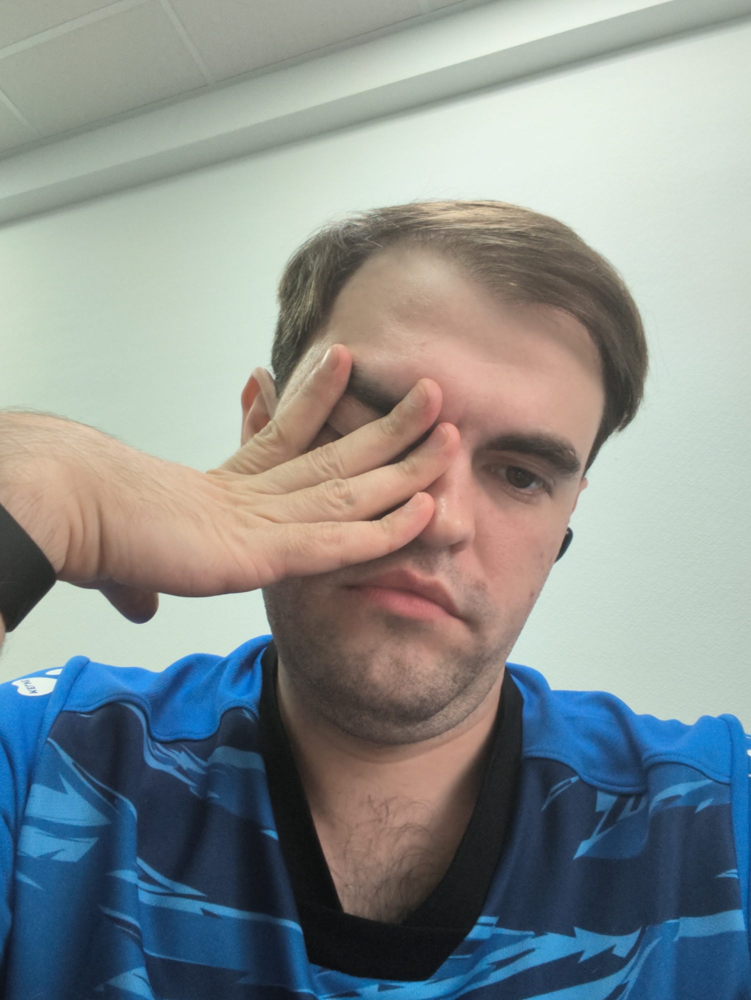 A man with light brown hair and a beard, wearing a blue and black patterned shirt, is captured from the chest up in a self-portrait. He is sitting, and his right hand is covering his right eye and part of his forehead. His left ear is visible, and he appears to be wearing a black wireless earbud in his left ear. The background is a plain, off-white wall with a white ceiling above. The lighting is bright and even.