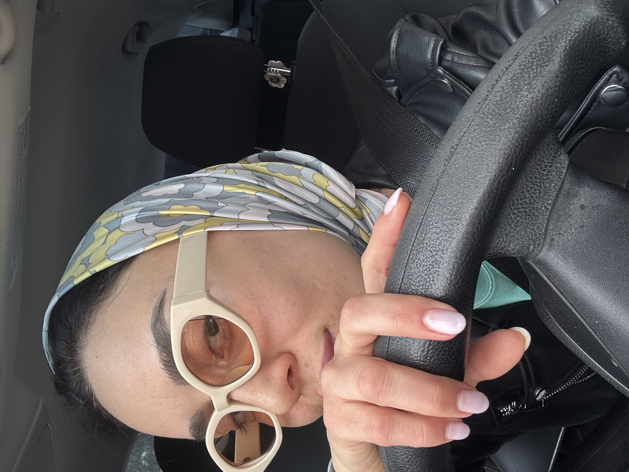 A close-up, rotated shot shows a woman in the driver's seat of a car, holding the steering wheel. She is wearing a patterned headscarf, featuring grey, yellow, and light blue cloud-like shapes, and large, beige-framed sunglasses with amber-tinted lenses. Her right hand, adorned with light pink nail polish, is gripping the black, textured steering wheel. Her mouth is slightly parted.
The car's interior is visible, with a black seatbelt crossing the frame and a black leather jacket or bag resting on a seat. A dark headrest is seen in the background, and a small, metallic decorative clip or brooch is attached to it. Parts of the car's light grey ceiling and door panel are also visible. Behind the steering wheel, a glimpse of a light blue garment or lining can be seen. The overall lighting suggests it is daytime. The scene is set in Tbilisi, Georgia.