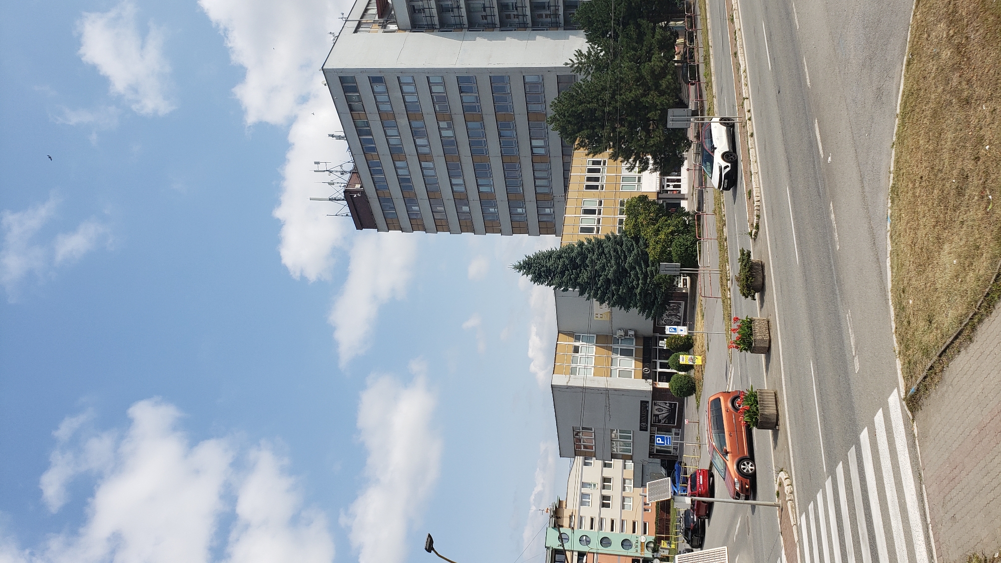 The image shows a street scene in Prešov, Slovakia. In the foreground, a crosswalk is visible on an asphalt road. On the right side of the road, a white car is driving. Further down the road, there are several buildings, including a tall, grey, multi-story building with many windows and several antennas on its roof. Next to it is a shorter, light-colored building with yellow accents and multiple windows. The middle ground features a large evergreen tree and several other trees and bushes. There are also planters with red flowers along the side of the road. The sky is blue with scattered white clouds. In the background, more buildings and trees are visible. The overall environment appears to be an urban setting on a clear day.