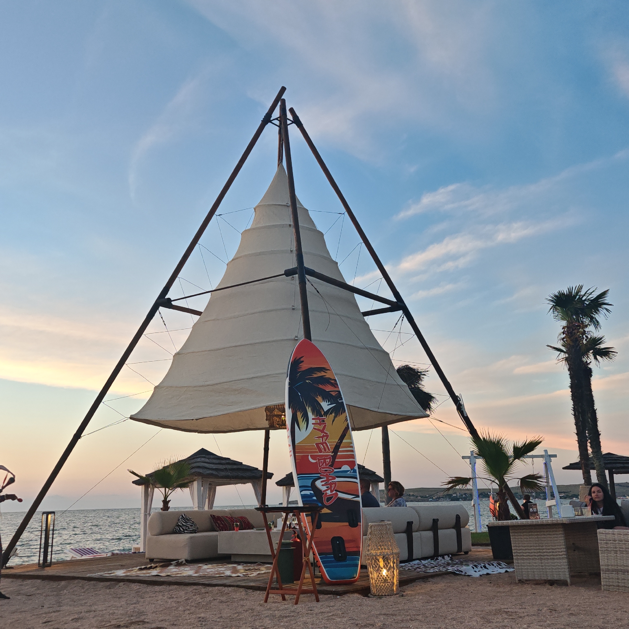 The scene is a beach resort at dusk, likely in Мысовское сельское поселение, Ukraine, with a large, cone-shaped white tent structure dominating the center. The tent is supported by a wooden frame and is illuminated from within by a warm light. A surfboard with a tropical design stands in front of the tent. In the background, there are several open-air seating areas with white couches and thatched-roof gazebos, positioned on a wooden deck overlooking the sea. Palm trees are visible to the right of the tent. Several people are seated at tables and on the couches, appearing relaxed. The sky is a soft blue with wispy clouds tinged with the colors of sunset. The overall atmosphere is serene and inviting, suggesting a resort or beachfront restaurant setting.