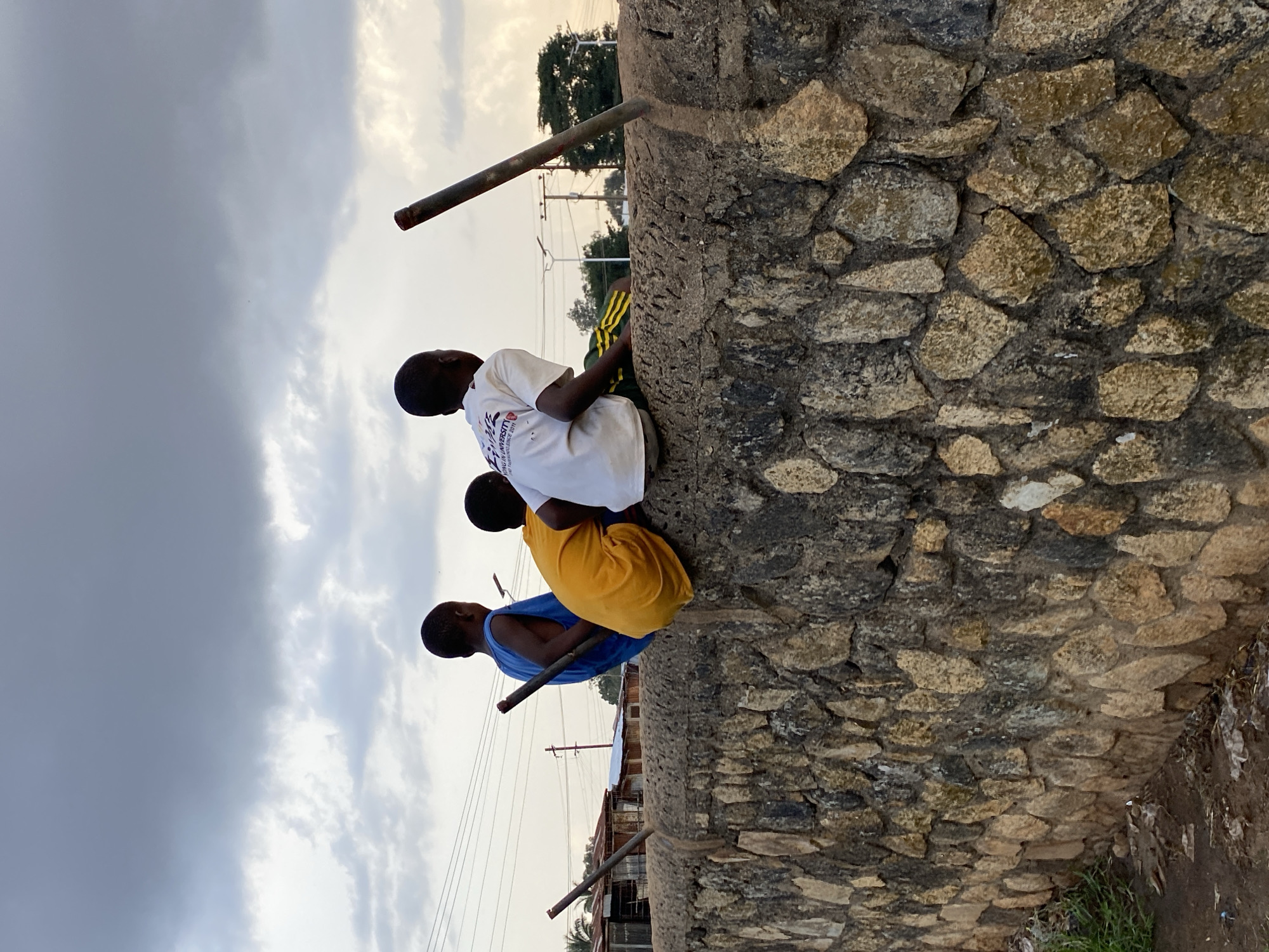 Three young boys are sitting on a rough, stone wall. The boy on the left wears a blue tank top and holds a stick. The middle boy wears a bright yellow t-shirt and is crouched low. The boy on the right wears a white t-shirt with writing on it and has yellow and green striped shorts. They are all facing away from the camera, looking towards the right side of the image. The sky above is overcast and gray. In the background, there are some buildings, power lines, and trees, suggesting an urban or semi-urban environment in Bukuru, Nigeria. The wall is made of irregularly shaped stones of varying colors, from light tan to dark gray. Some debris is visible on the ground at the base of the wall. Two metal poles are embedded vertically into the top of the wall, one behind the boy in blue and another to the right of the boy in white.