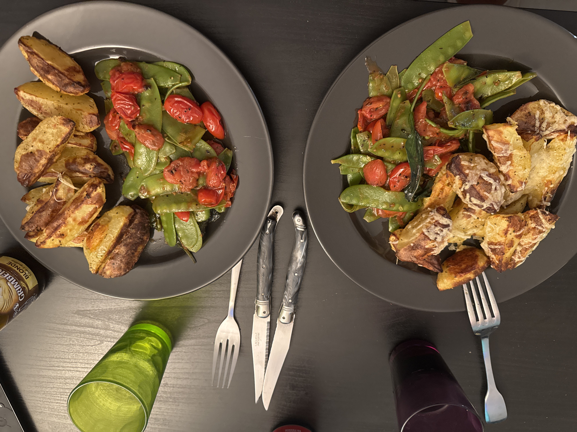 The image depicts a meal for two, likely in a private residence. The setting is a dark surface, possibly a table. Two grey plates are present, each containing roasted potatoes and a mixture of sauteed snap peas and roasted cherry tomatoes. Utensils, including forks and steak knives, are positioned between the plates. A bottle of Grimbergen Blonde beer is visible on the left, and two glasses (one green, one purple) are also on the table. No people are visible. The setting lacks identifiable markers suggesting a specific location in Toulouse, France.