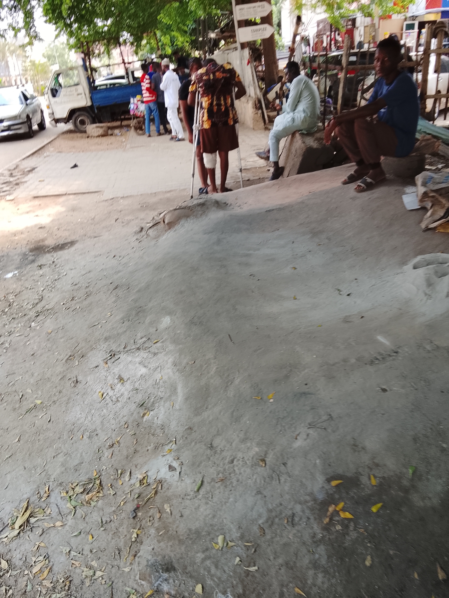 In Abuja, Nigeria, a scene unfolds on a dusty street with a white truck parked on the left. Several people are present, including a man with a crutch standing near a food stall. Another man is seated on a raised concrete structure, facing away from the camera. The environment is characterized by dry earth, scattered leaves, and the presence of debris. The time of day appears to be daytime, with sunlight casting shadows.