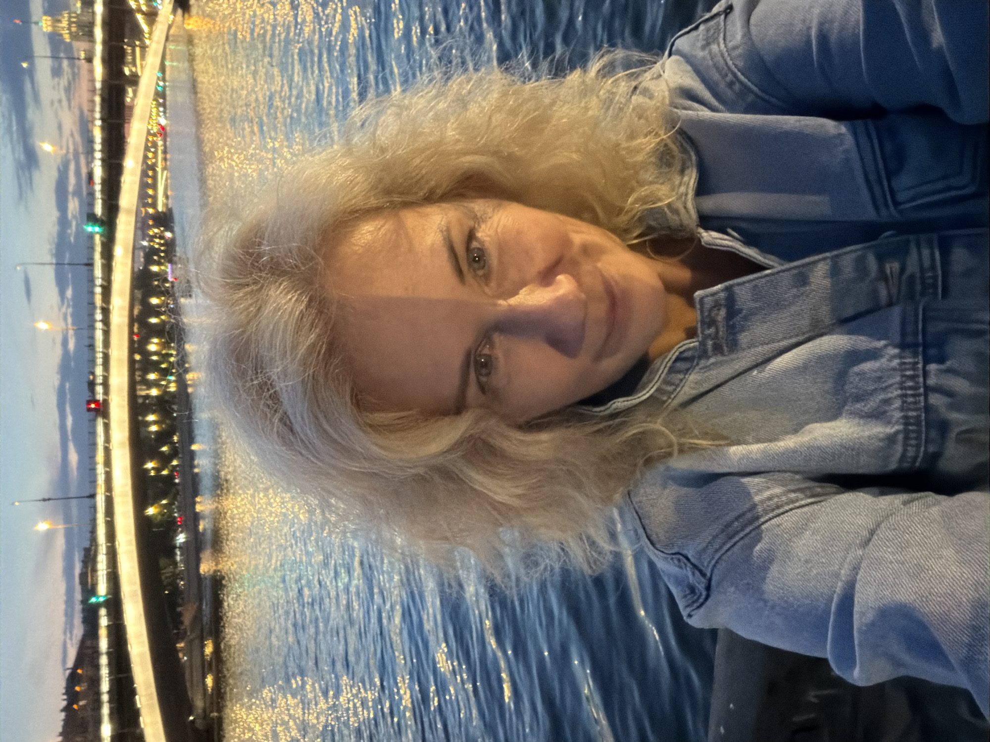 In an outdoor, medium shot at dusk, a woman with blonde, curly hair takes a selfie in front of a river and a bridge. The woman is looking at the camera and smiling, and she is wearing a light blue denim jacket. The river is dark blue with reflections of lights from the city. The bridge in the background is illuminated with yellow lights, and there are lights from buildings on the far bank of the river. The sky is a pale blue with some clouds. The scene appears to be in Moscow, Russia, possibly along the Moskva River.