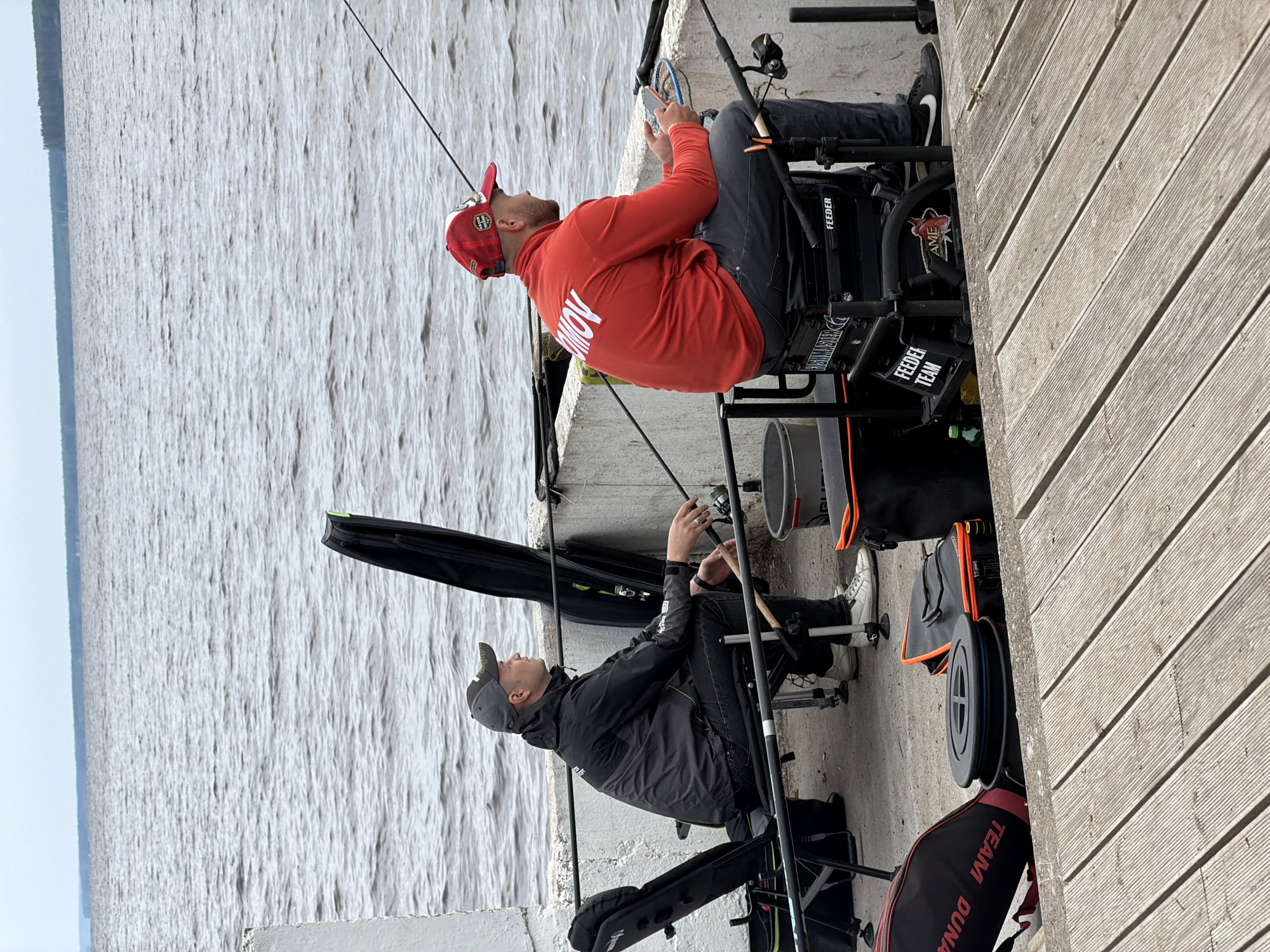 Two men are engaged in fishing activities from a concrete and wooden pier or embankment next to a large body of water, likely a river or reservoir in Chaikovsky, Russia. The image is taken during the daytime under what appears to be an overcast sky, providing soft, even lighting.
Both men are seated on specialized fishing chairs or platforms, equipped with multiple fishing rods and other gear. The man in the foreground, wearing a red baseball cap and a red long-sleeved shirt with the partial text "ROMOV" on the back, is looking at a smartphone. He has dark pants and Nike sneakers. His fishing setup includes a black chair with "FEEDER TEAM" and "FEEDER" visible, and a cooler or bucket nearby.
Behind him, another man in a dark baseball cap, dark jacket, and dark pants is looking out towards the water, actively tending to a fishing rod. His setup is similar, with a black fishing chair and various fishing accessories. Near him, a black bag with "TEAM DUNA" in red lettering is visible, along with other equipment. The water surface shows light ripples. The overall scene depicts a calm day for competitive or leisure feeder fishing.
