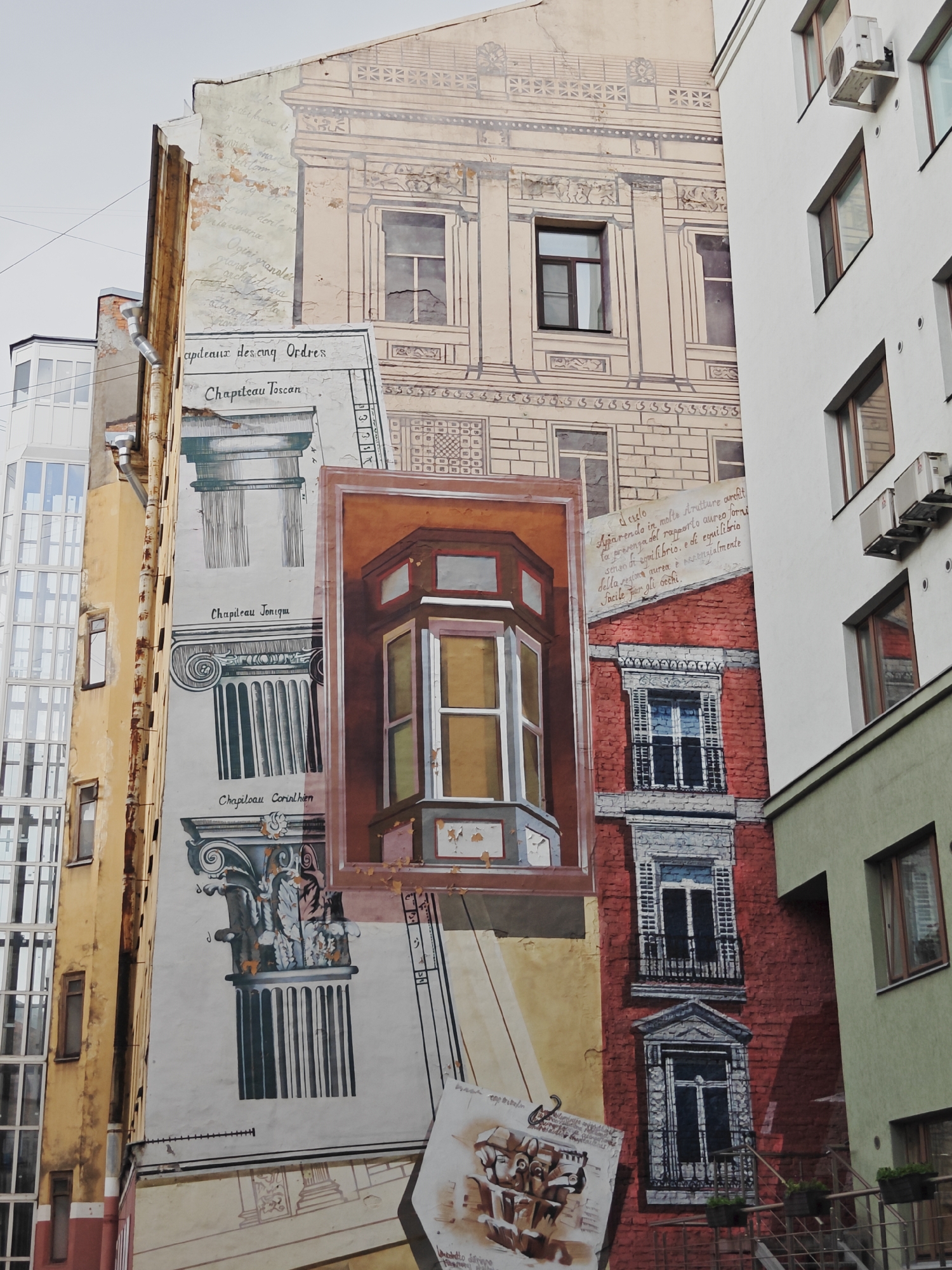 The image captures a section of a building in Saint Petersburg, Russia, adorned with a large, detailed mural. The mural depicts architectural elements, including classical columns labeled "Chapiteau Toscan," "Chapiteau Ionique," and "Chapiteau Corinthien," rendered in black and white line drawings. Interspersed with these are sections of painted architecture, featuring windows and balconies, some in a reddish brick facade, and others in a lighter, classical style. A prominent element is a painted bay window with colorful glass panes. The mural also includes handwritten text in what appears to be French and Italian, adding to the artistic composition. The mural covers the facade of an older building, with portions of peeling paint and weathered surfaces visible. Adjacent to this muraled building are other structures: a modern building with extensive glass windows on the left, and a white building with multiple windows and air conditioning units on the right, partially obscuring the mural. The overall scene is an urban environment, with the sky visible in the upper portion of the image. No people are visible in the frame.