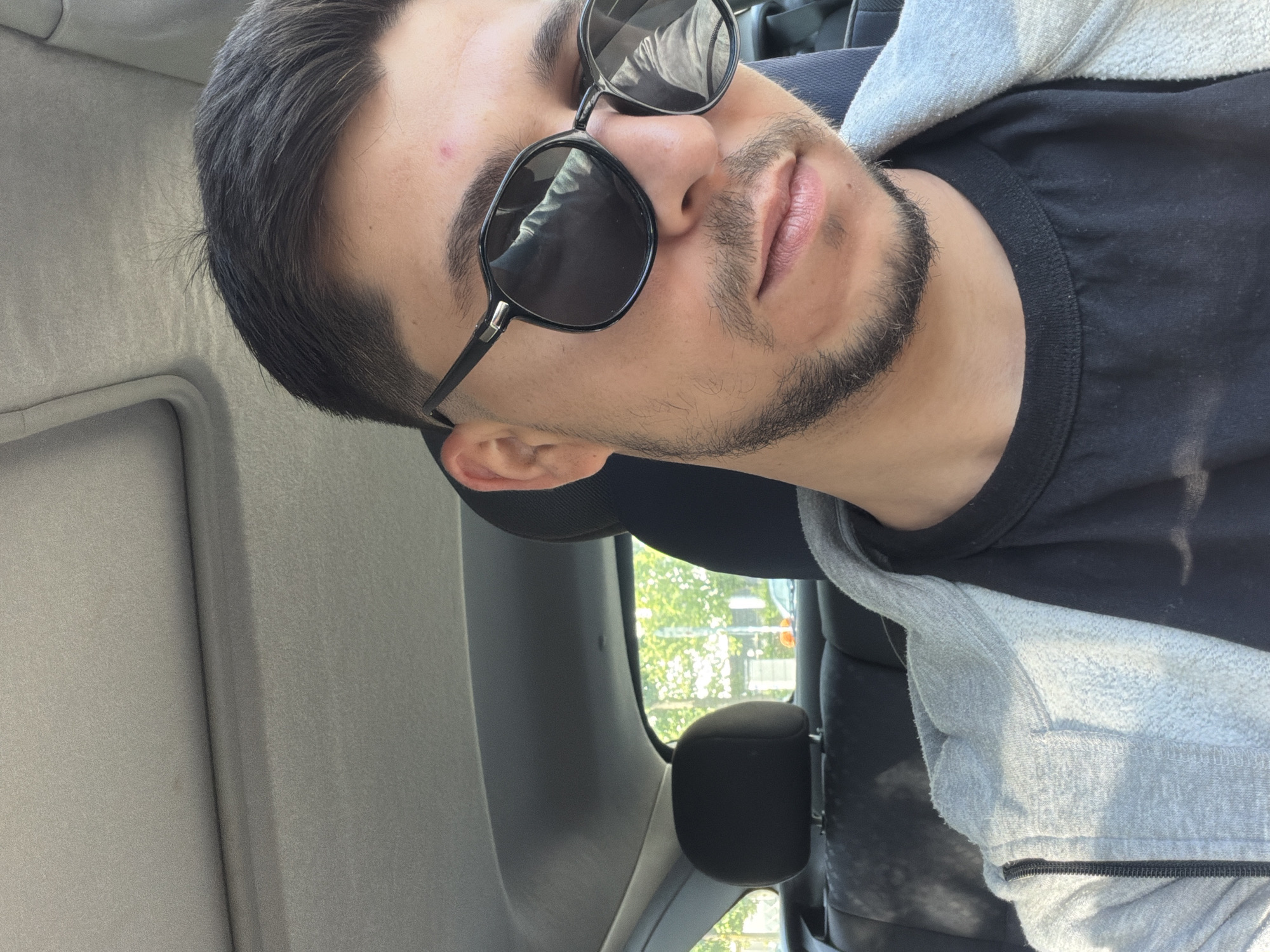 A close-up shot looking up at a man in sunglasses. He is seated in a car, visible by the interior details such as the headrest and sun visor. Outside the car window, trees can be seen, indicating a location with greenery. The man has dark hair and a beard. He wears a black t-shirt and a gray hoodie. The city of Gütersloh, Germany, cannot be confirmed from the image.
