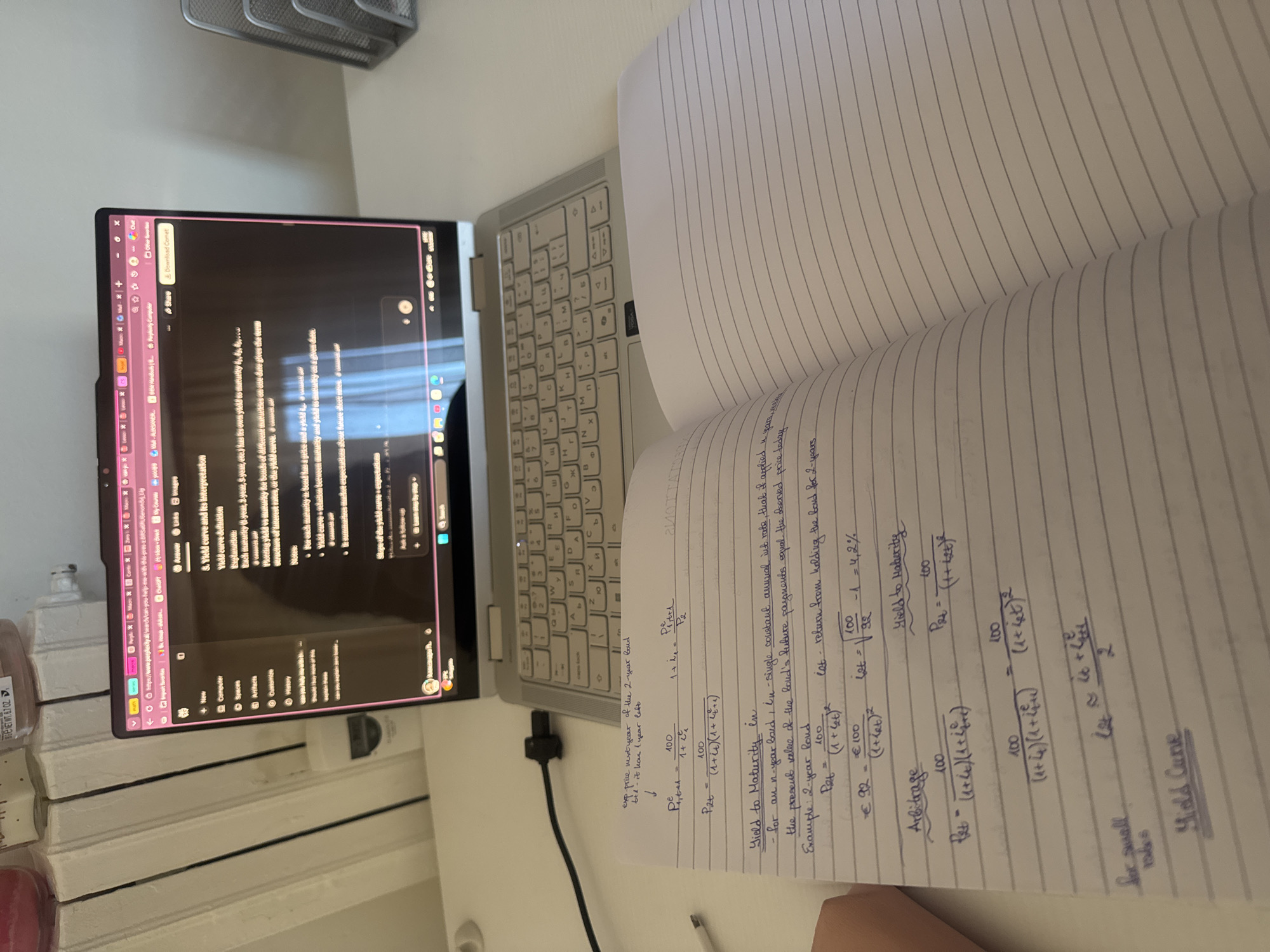 The image shows a desk with a laptop, a notebook, and some desk accessories. The laptop screen displays information about "Yield Curve and its interpretation," suggesting a study or work context related to finance or economics. Open to a page of handwritten notes, the notebook contains mathematical formulas and calculations pertaining to "Yield to Maturity," "Arbitrage," and "Yield Curve." The overall setting implies a workspace or study area, possibly in a home environment. The image does not provide information to determine a location in Milan, Italy.