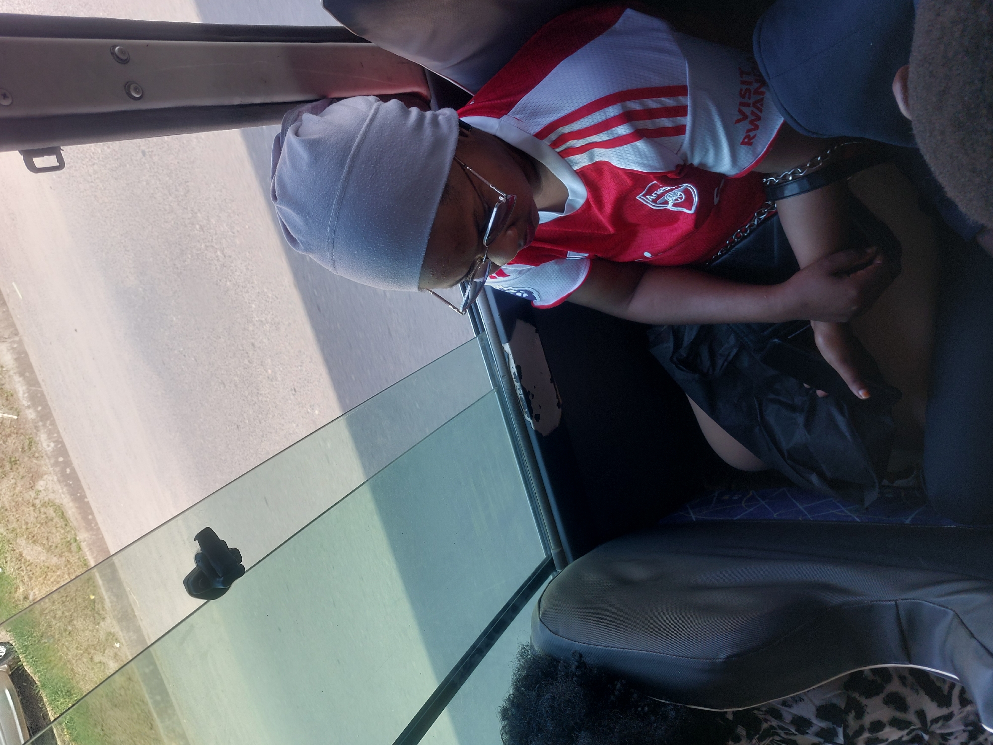 A person wearing an Arsenal football jersey, grey head covering, and glasses is seated in what appears to be a vehicle. The person has a thick metal collar secured around their neck with a chain. They are holding the chain. The vehicle has a window beside the person, showing a road and some greenery outside. The person is wearing a black garment on the bottom. Parts of vehicle seats are visible, with the bottom corner showing an area with an animal print design. There is no definitive evidence within the image to confirm the location is Dar es Salaam, Tanzania.
