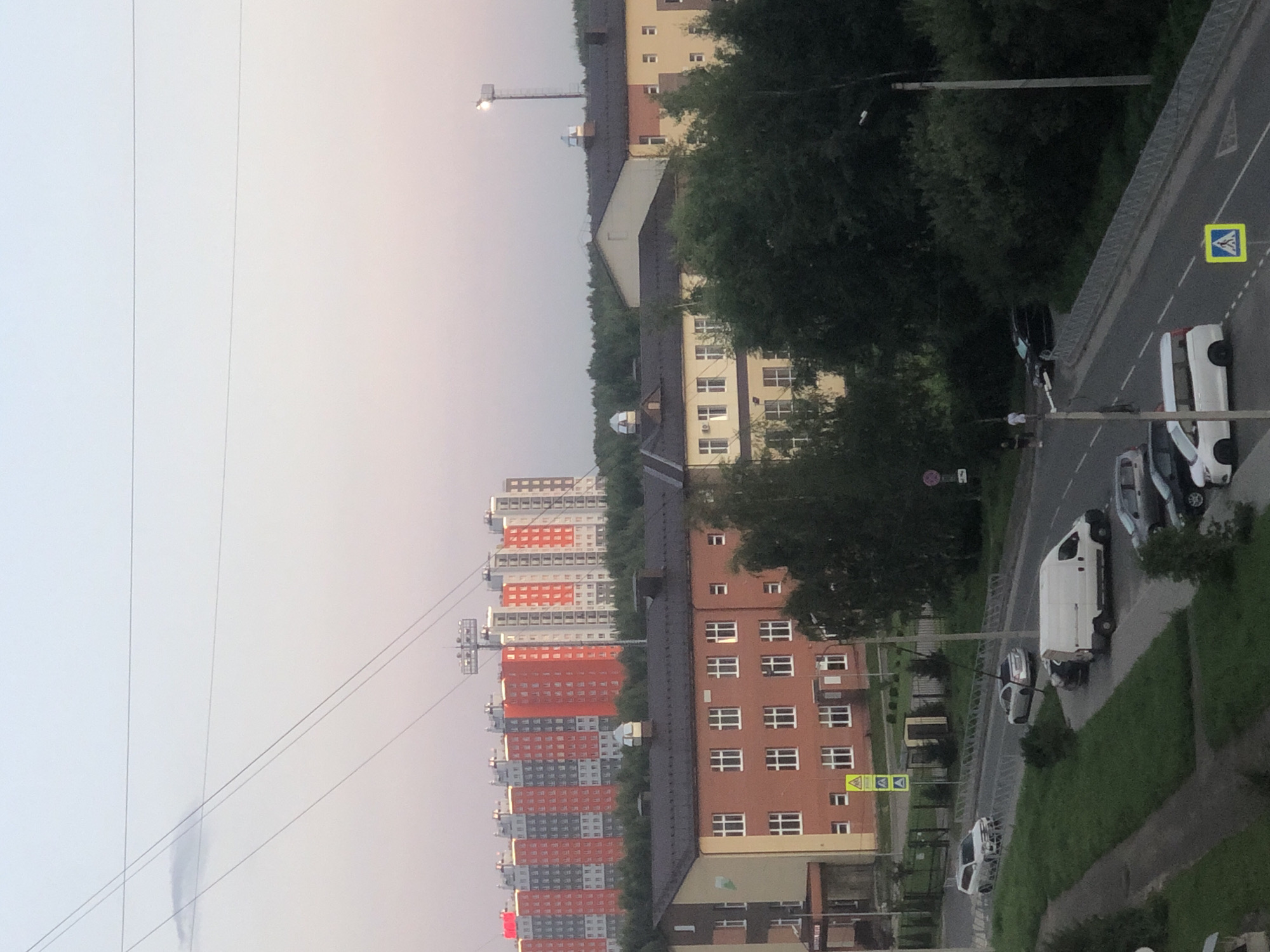 The image depicts an urban scene in Rusanovo, Russia, bathed in the soft light of dusk. In the background, a row of vibrantly colored apartment buildings, some featuring red and orange facades, stand tall against the pale sky, punctuated by overhead power lines and a distant street lamp. Closer to the foreground, a low-rise brick building with a dark roof and numerous windows is visible. A paved road runs through the midground and foreground, with several vehicles present. A white van is parked on the side of the road, and a silver sedan and a white SUV are driving on the road. Two pedestrians are walking on a sidewalk near the road. The environment is characterized by a mix of urban architecture and lush green trees.