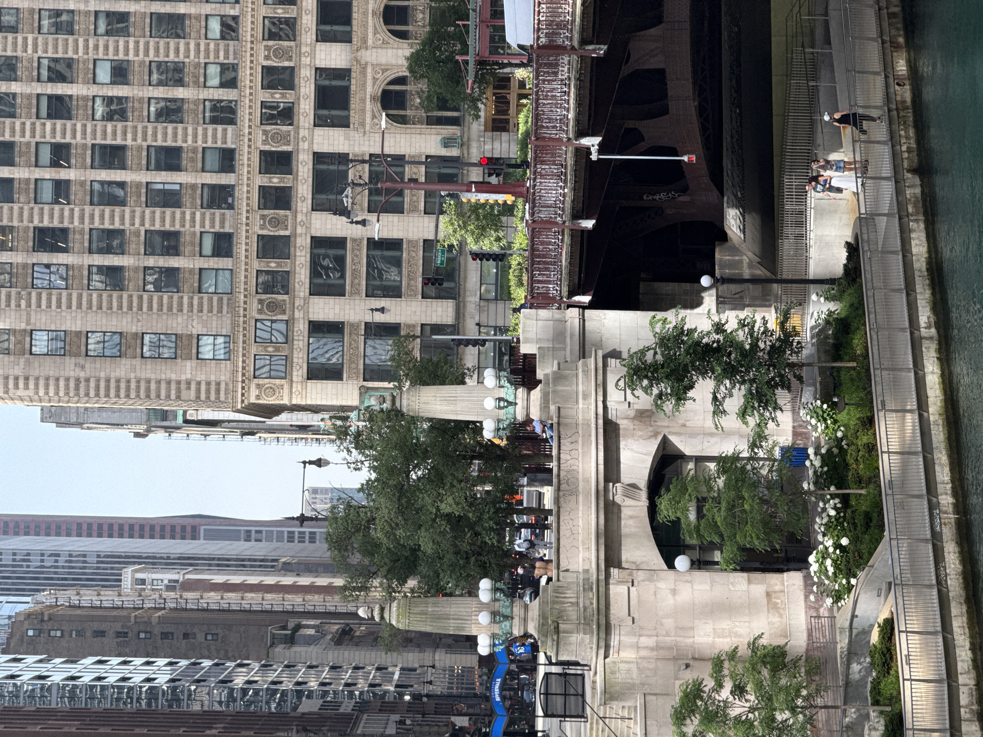 The image depicts a view of the Chicago River in Chicago, Illinois, USA. In the foreground, the river is visible with its greenish-blue water. A concrete embankment lines the river, topped by a metal railing. Beyond the railing, a paved walkway curves gently. On this walkway, three individuals are present: two women appear to be conversing, and a woman wearing a white hat stands facing away from the camera.
Behind the walkway, a section of a stone bridge pier is visible, featuring arched openings and decorative stonework. Further back, a tree-lined area with white flowers adds a touch of greenery. Several trees stand on a lower level, casting shadows.
Above the bridge pier, a multi-story building with a light-colored stone facade and numerous windows dominates the midground. A traffic light with red and yellow signals is visible on the left side of the frame. A street sign indicating "Wabash Ave" is also present. A dark red bridge with a decorative metal railing spans across the river in the midground.
In the background, a dense urban landscape of tall buildings with various architectural styles and materials rises into a hazy sky. Some buildings have a grid-like pattern of windows, while others feature decorative elements. The overall scene suggests a sunny day in a bustling urban environment.