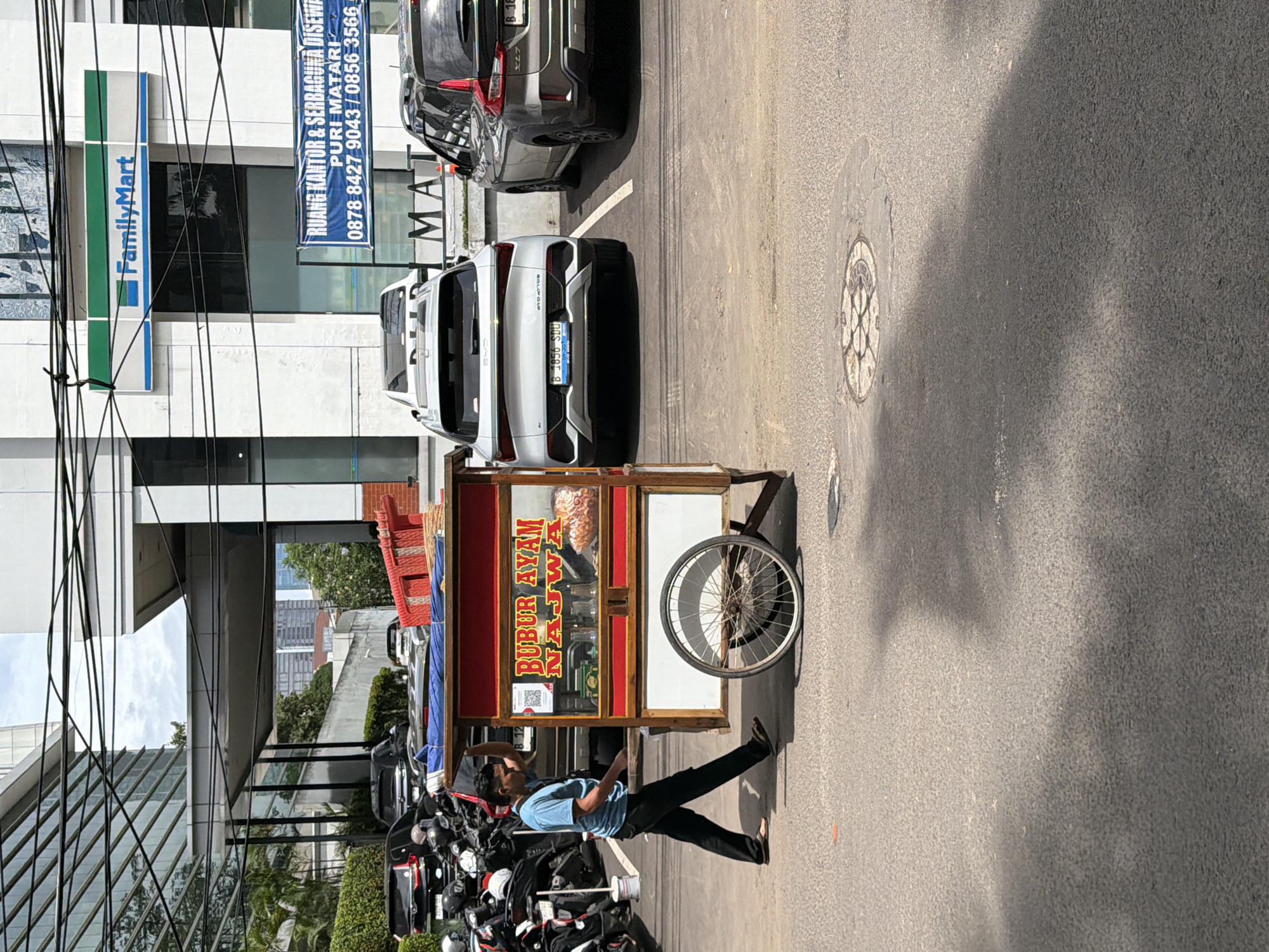 The image captures a street scene in South Jakarta, Indonesia. A vendor pushes a "Bubur Ayam Najwa" food cart along the side of the road. Behind the vendor, several parked motorcycles are visible. Cars are parked in front of an office building. Signage on the building indicates "FamilyMart" and "RUANG KANTOR & SERBAGUNA DISEWA PURI MATARI" with associated phone numbers. A manhole cover is visible on the street.
