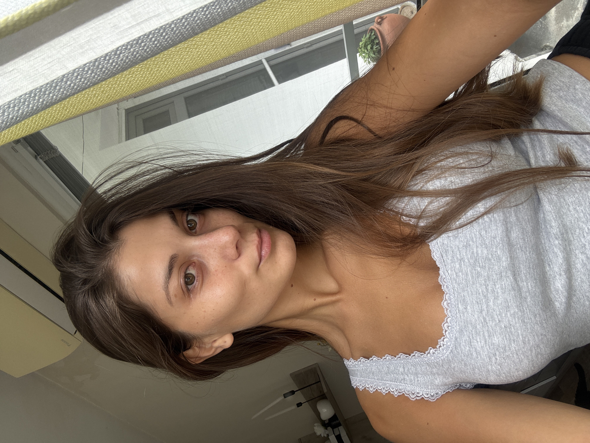 A woman is in an indoor environment in Burgas, Bulgaria. She has brown hair and brown eyes and is wearing a gray tank top with white lace trim. She is near a window, with striped blinds and a view of buildings outside. There is a potted plant on the window sill.
