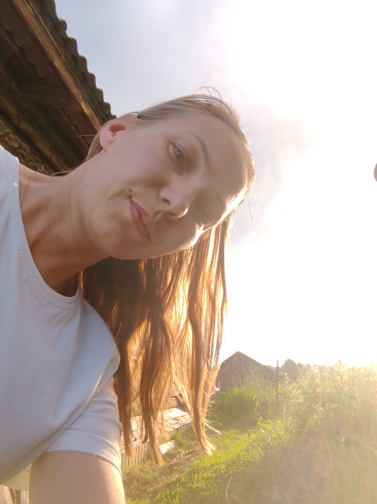 A woman, identified as potentially being in Chuchlam, Russia, is captured in a low-angle, selfie-style photograph. She is wearing a white t-shirt and has long, auburn hair that is illuminated by the sun, creating a halo effect. The woman's head is tilted, and she is looking towards the camera with a slight smile. Her skin appears fair, with some freckles visible on her cheeks and forehead. A thin, gold-colored necklace is visible around her neck.
The background suggests an outdoor, rural setting. To the upper left, a portion of a rustic wooden structure, possibly a roof or awning, is visible. The sky is bright and washed out by sunlight, indicating it is daytime, likely late afternoon or early evening due to the warm glow. To the right of the woman, a field of tall green grass and vegetation is present, with a small wooden structure, possibly a shed or outbuilding, visible in the distance. The sunlight is creating a strong lens flare, partially obscuring details in the background. The overall impression is one of a casual, sun-drenched moment outdoors.