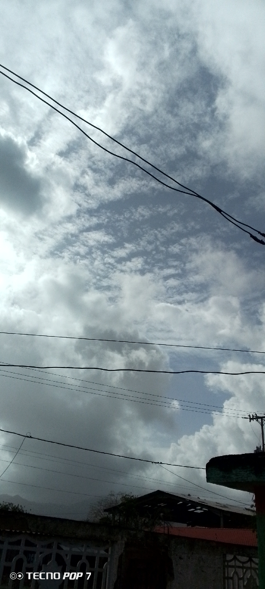 The image shows a cloudy sky above a residential area in Altagracia, Venezuela. Several black electrical wires stretch across the frame, disrupting the view of the sky. Beneath the wires, the tops of buildings and trees are visible. The structures appear to be modest homes, with a mix of roof styles and materials. The overall environment suggests a typical residential neighborhood in the town. The sky is predominantly covered with grey and white clouds, with patches of blue visible in between. The lighting is subdued, indicating an overcast day. There are no people visible in the scene. The primary subjects are the sky, electrical wires, and the rooftops of the houses. The image appears to have been taken from a ground-level perspective.