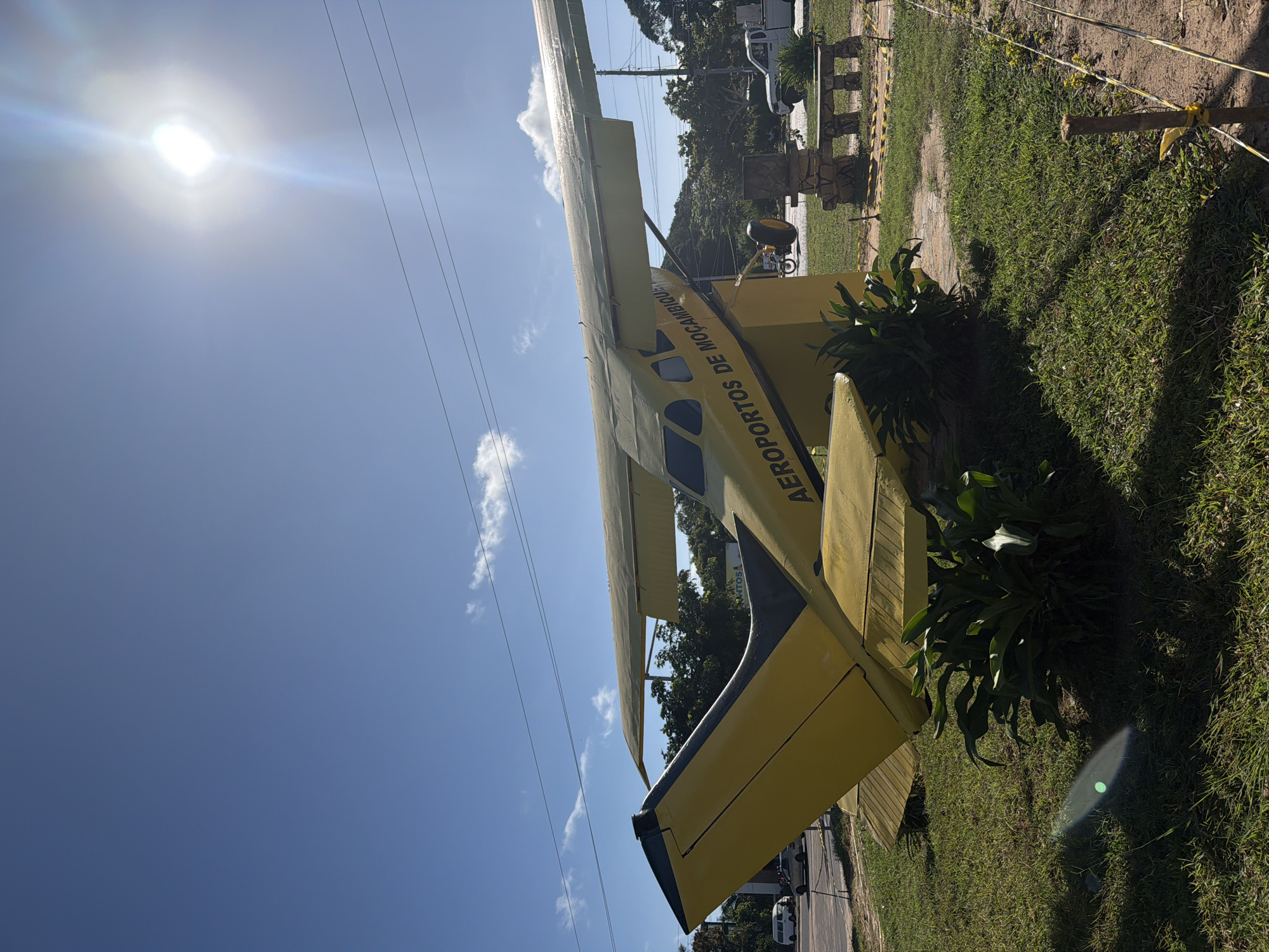 The image displays a bright yellow single-engine aircraft, likely a bush plane, prominently positioned outdoors under a clear blue sky. The airplane is a static display, situated on a grassy patch of ground next to what appears to be a road or paved area. The sun is very bright, casting a strong glare and lens flare in the upper left portion of the sky.
The aircraft itself is yellow with black accents on its tail section and wings. The text "AEROPORTOS DE MOÇAMBIQUE" is clearly visible in blue lettering along the fuselage, indicating it belongs to "Airports of Mozambique." The plane appears to be supported by a yellow structure, possibly a stand, matching the plane's color.
In the background, beyond the aircraft, there are lush green trees and some power lines stretching across the sky. A white utility truck and other vehicles are partially visible in the distance, suggesting a road or parking area nearby. On the ground, there are patches of well-maintained grass and some darker green foliage, possibly shrubs, around the base of the aircraft display. Several concrete pillars or decorative structures are also visible in the background, further suggesting a public display or monument. The overall scene indicates a sunny day with minimal cloud cover.