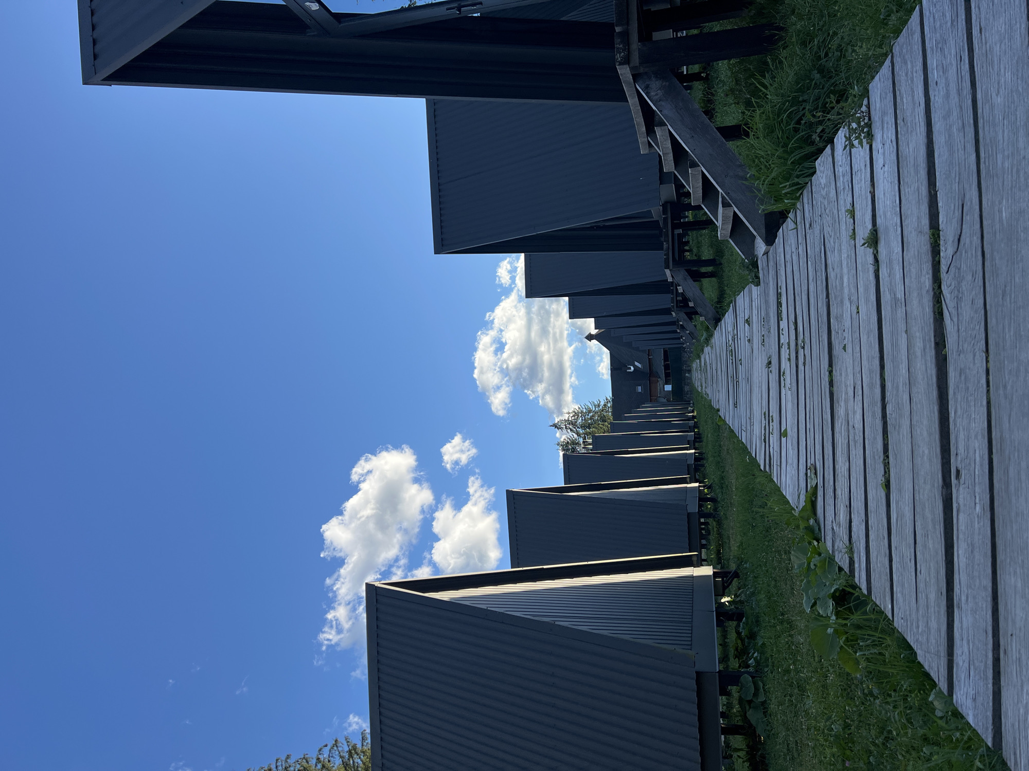 The image depicts a row of modern, dark gray A-frame cabins set on stilts, likely part of a resort or accommodation complex in Vorokhta, Ukraine. A wooden boardwalk stretches away from the viewer, leading towards a central building in the distance, also appearing to be part of the same complex. The cabins are lined up symmetrically along the boardwalk, with stairs leading up to some of them. The environment is natural, with green grass and some foliage around the base of the cabins. The sky is a clear blue with a few scattered white clouds, suggesting fair weather. There are no people visible in the image. The overall scene is one of ordered, modern construction integrated into a natural, possibly mountainous, setting.