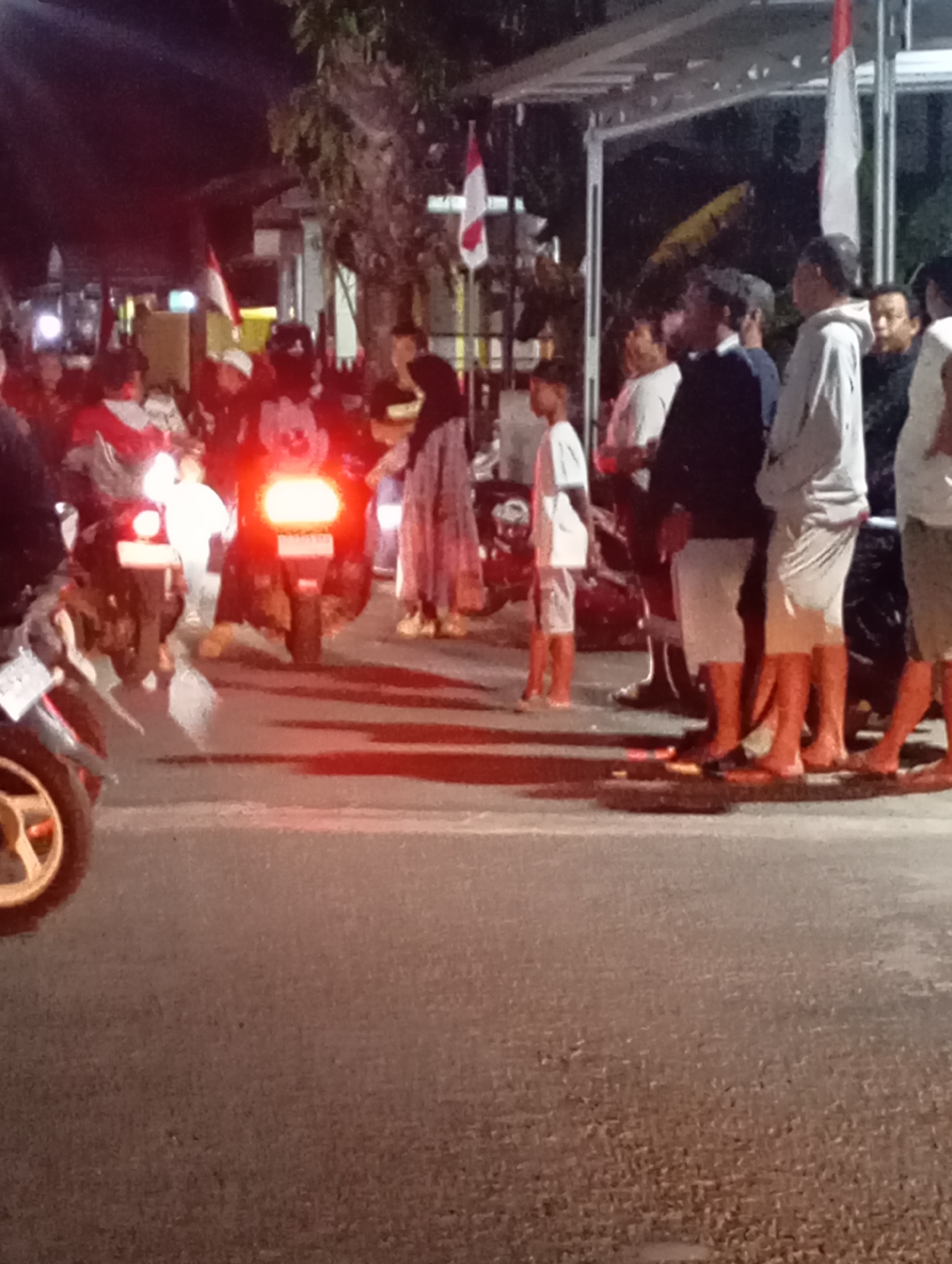 The image depicts a nighttime street scene in Pacitan, Indonesia, likely near a commercial or gathering area. A group of people, a mix of adults and possibly children, are present. Several motorcycles are parked, with their rear lights illuminated, casting red and orange glows on the asphalt. One motorcycle in the foreground on the left has its front wheel visible.
The individuals are casually dressed. Some are wearing t-shirts, shorts, and flip-flops, common attire for warm weather and informal settings. One woman on the right is wearing a headscarf and a long, patterned dress. Several Indonesian flags, red and white, are visible, suggesting a national event or celebration. The background shows a simple structure with a white roof, possibly a shop awning or a small building. Trees with dense foliage are also present, contributing to the urban outdoor environment. The overall impression is one of a community gathering or event taking place after dark, with a focus on motorcycles and patriotic symbols. The lighting is dim, with the primary illumination coming from the motorcycle lights and some unseen overhead sources.