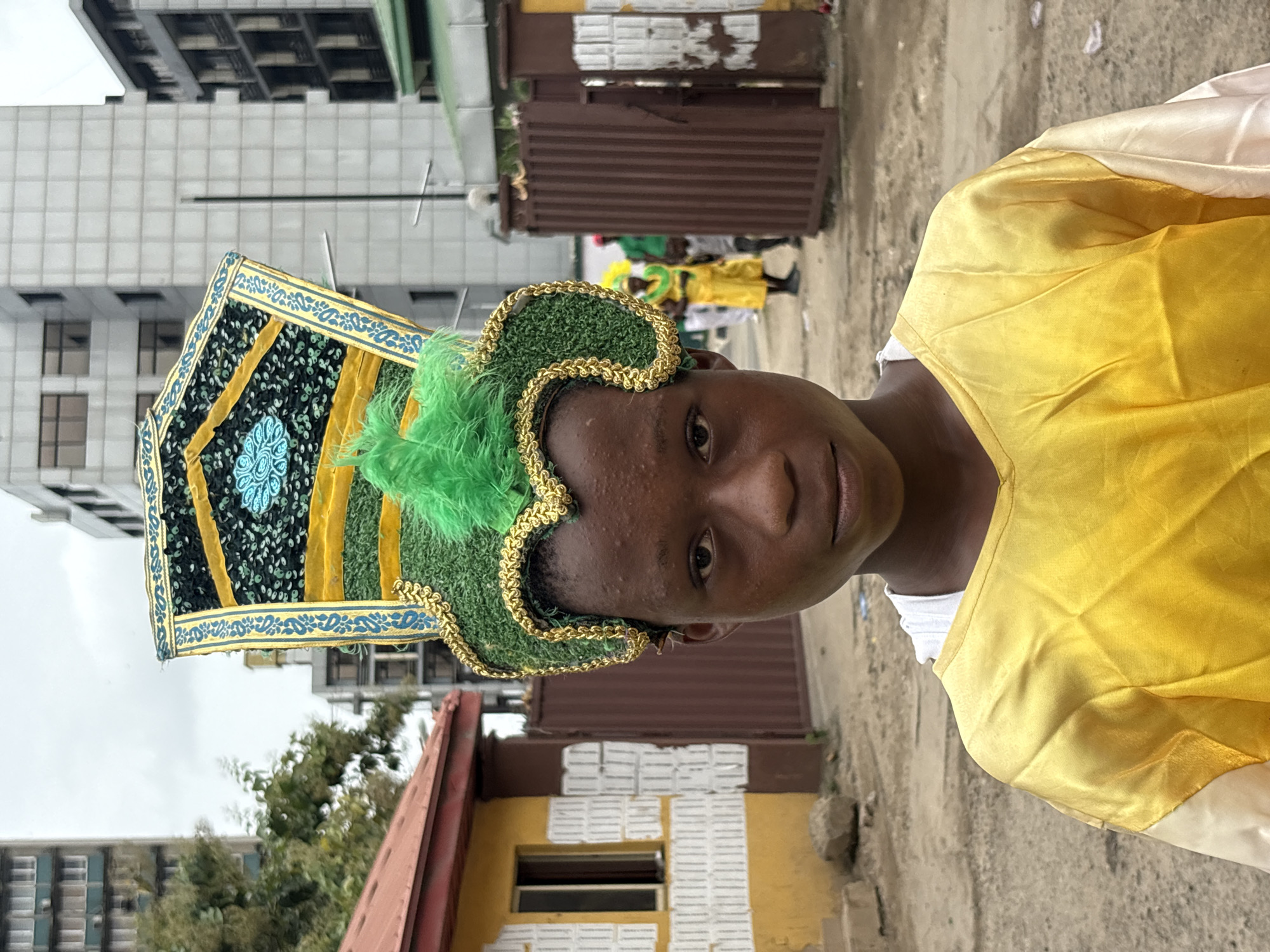 A close-up, outdoor shot, oriented vertically, features a young person adorned in vibrant traditional attire, likely participating in a cultural celebration or event. The individual, with brown skin, is positioned at the forefront, their face directed towards the camera. They wear a yellow, satin-like tunic and a green headpiece embellished with green feather-like accents, edged with gold. A large, square, decorated hat stands atop the headpiece.
In the blurry background, other people are seen in similar attire. A dilapidated building with a red roof is adjacent to a yellow building, which is partially obscured by the person in the foreground. Further behind, more people in colorful costumes are indistinctly visible.
The setting appears to be an urban environment, with modern buildings towering in the background. This is likely Lagos, Nigeria, given the style of clothing and architecture present. The ground is a mixture of dirt and pavement. The shot is candid and unposed, capturing a moment in a public space.