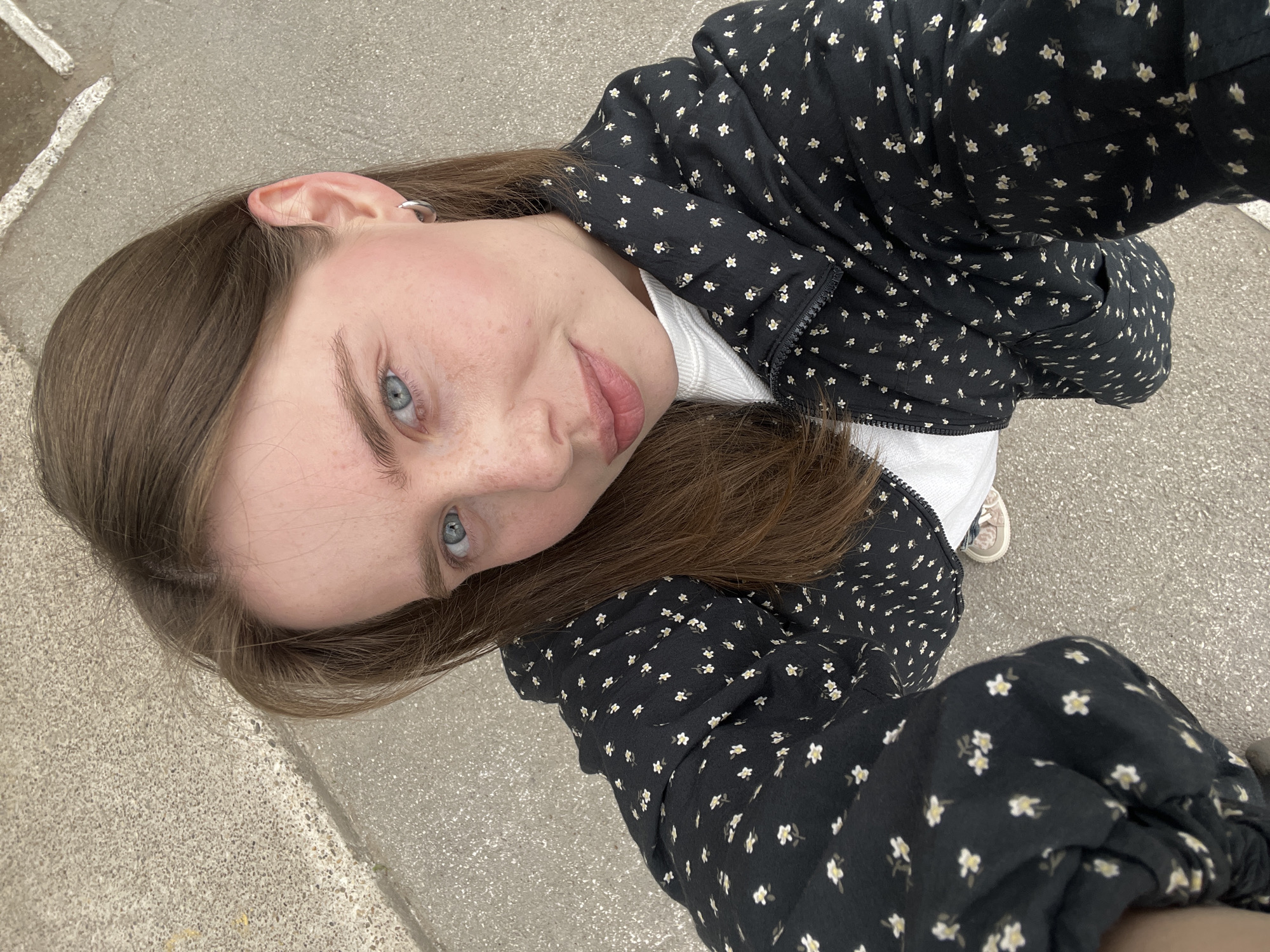 A young individual, likely female, is captured in a close-up, upward-angled self-portrait. She has long, straight brown hair that frames her face, clear blue eyes, and visible freckles across her nose and cheeks, which show a subtle flush of pink. She wears a black jacket adorned with a repeating pattern of small white and yellow flowers, layered over a white top. A small silver hoop earring is visible in her right ear.
The setting is outdoors, with a uniform, light grey paved surface forming the background, suggesting a sidewalk, pathway, or similar urban/suburban ground in Chaikovsky, Russia. The lighting indicates it is daytime, and the weather appears clear. The individual is looking directly into the camera with a soft, neutral expression that carries a hint of a gentle smile. A portion of a shoe, possibly a sneaker, with light-colored laces, is visible near the bottom right, suggesting the individual is standing or sitting on the ground. There is no visible text in the image.