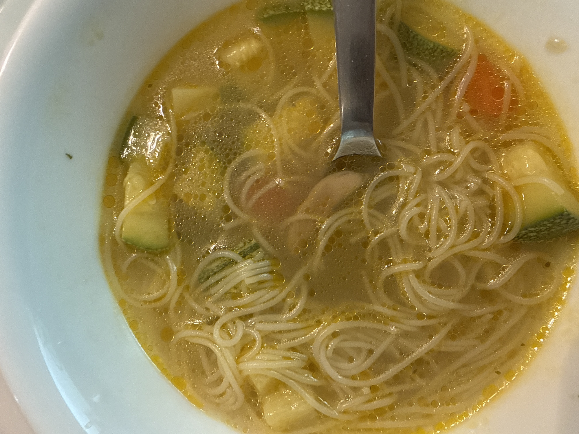 A top-down shot depicts a bowl of noodle soup, possibly chicken noodle soup, in a white bowl. The soup contains thin noodles, visible vegetables (likely zucchini and a piece of carrot), and what seems to be pieces of chicken. A metal spoon is partially submerged in the soup. The broth has an oily surface with visible bubbles. The scene is indoors with possibly natural lighting. There is no information to determine its location in San Bernardino Tlaxcalancingo, Mexico, from the image alone.