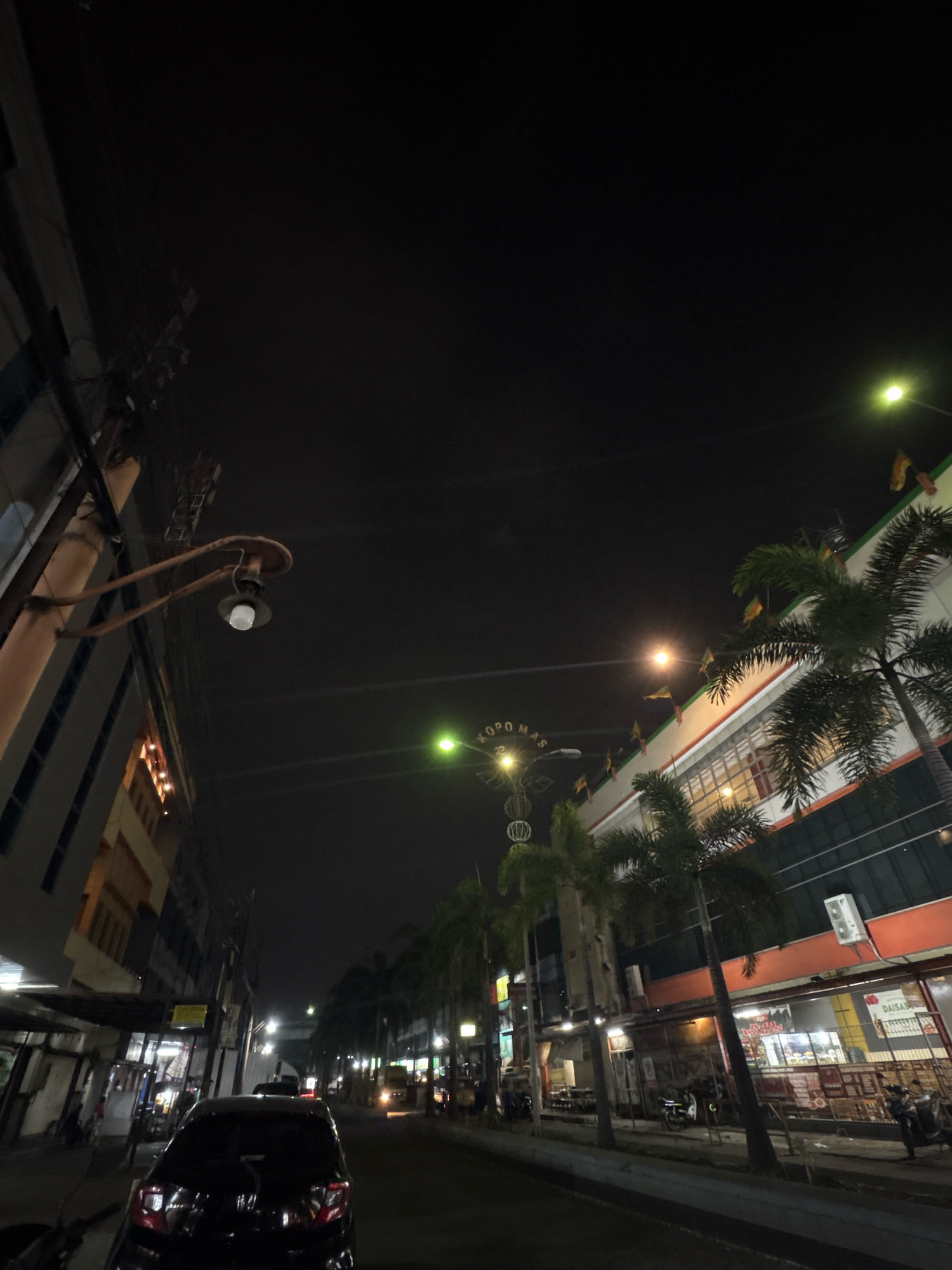 The image depicts a street scene at night in Bandung City, Indonesia, likely in a commercial area. Tall buildings with illuminated windows line the left side of the street. On the right, a multi-story building with orange and white trim is visible, featuring palm trees in front of it. Several streetlights illuminate the scene, casting a yellowish glow. The street itself is paved, with a dark car parked in the foreground. The sky is dark, indicating it is nighttime. The overall impression is of a bustling urban environment after dark, with a mix of modern and older architecture.