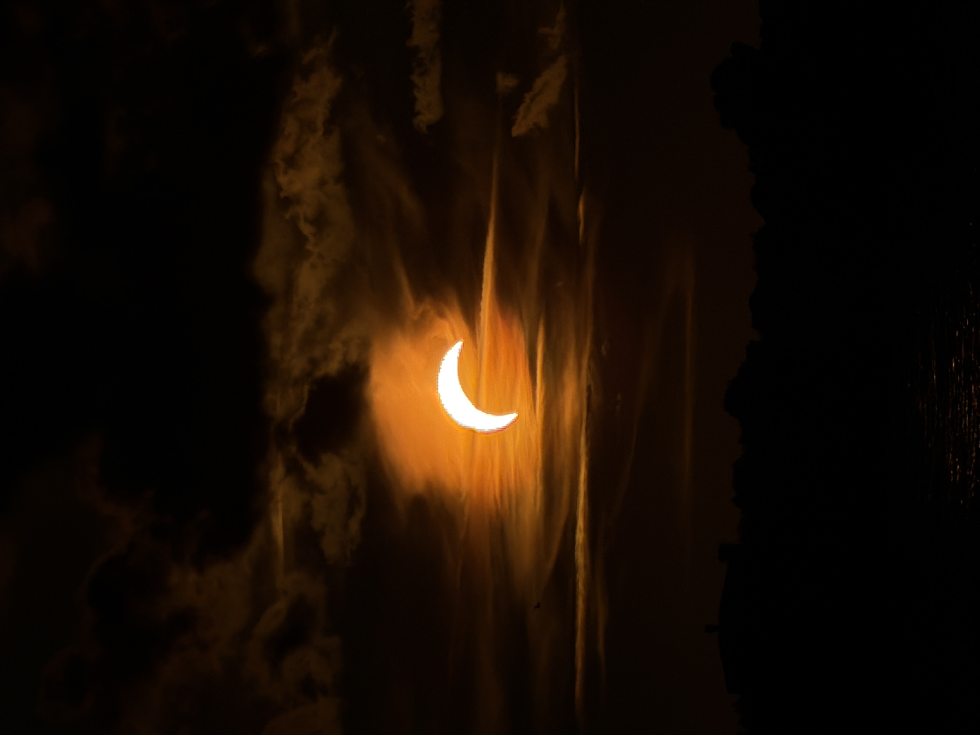 The image shows a partial solar eclipse over the Daugava River in Riga, Latvia. The sun is partially obscured by the moon, creating a crescent shape. The sky is filled with dark, ominous clouds, tinged with orange and yellow from the sun's light. In the foreground, the silhouette of trees and buildings lines the riverbank. The water in the foreground reflects the dim light of the sky. A small bird is visible in the sky to the left of the sun.