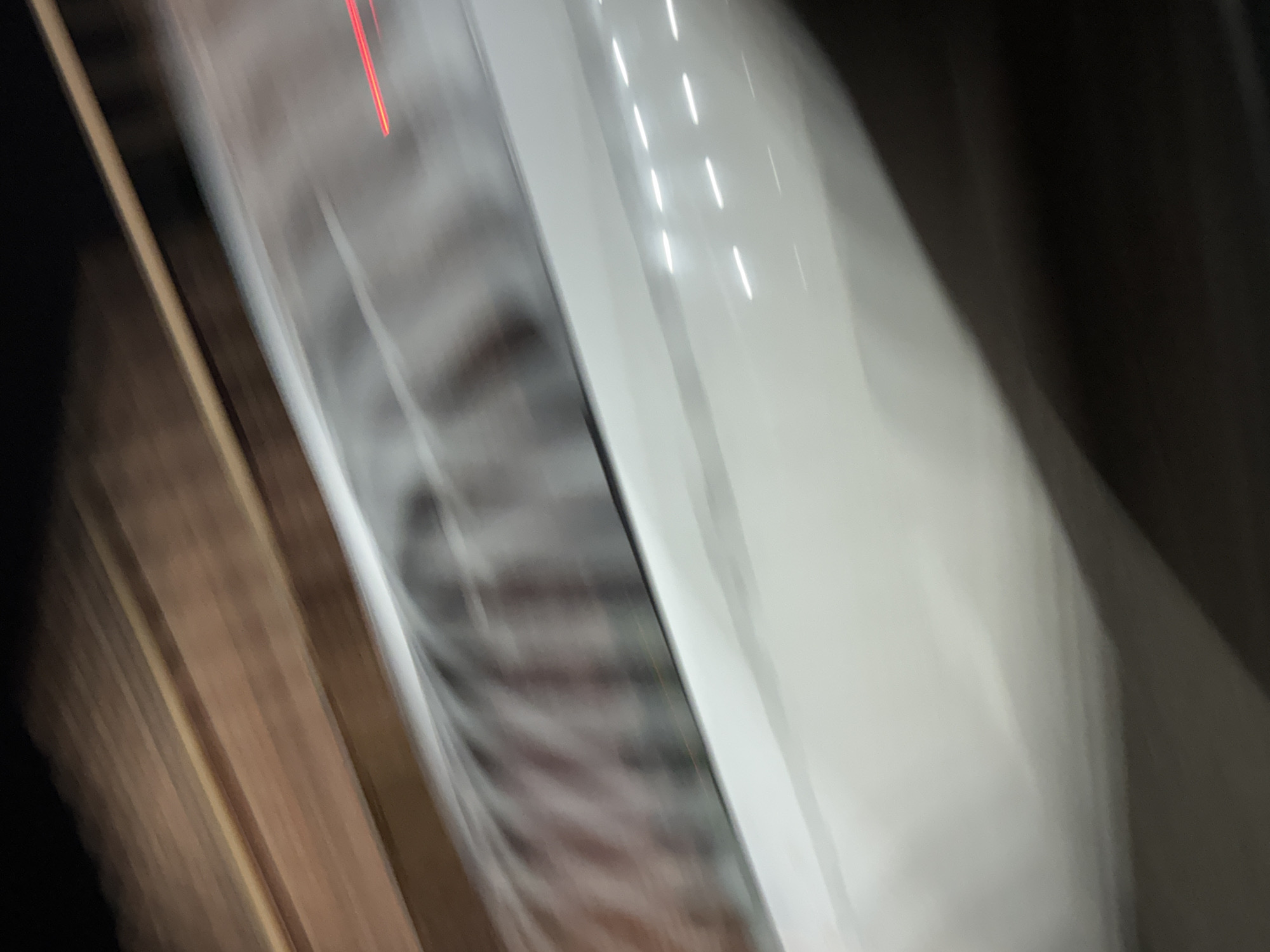 This is a blurry, motion-streaked photograph of a vehicle in motion, likely taken at night or in low light conditions. The foreground shows the white exterior of the vehicle, with several horizontal white lights visible, possibly interior lights reflecting or illuminated elements on the vehicle's surface. Above the white surface, there is a blurry depiction of a railing or some textured material, with vertical elements creating a ribbed pattern. Further up, a dark brown, textured surface is visible, with horizontal lines suggesting wooden planks or a similar material. A faint red streak is visible on the right side of the image, indicating a light source from another moving object. Due to the motion blur and lack of discernible landmarks or specific architectural features, it is not possible to determine the exact location within Grozny, Russia.
