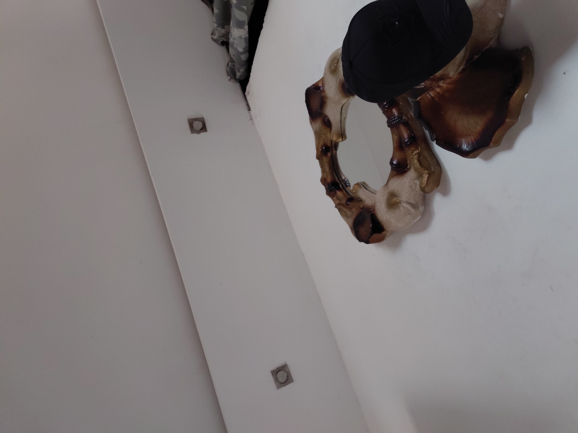 An angled shot captures an indoor scene, likely a room in Santa Ana, Venezuela, dominated by white walls and/or a ceiling. The perspective looks upwards and diagonally across the space.
The primary subject is an ornate, decorative mirror positioned on the white wall. Its frame appears to be crafted from a material resembling bone, wood, or a composite, with varying shades of beige, cream, and dark brown, suggesting an aged or organic aesthetic. A black hat, possibly a fedora or trilby style, is placed on top of the mirror frame, resting partially on the mirror's surface.
Further along the white surface, two small, square-shaped recessed light fixtures or sensors are visible. In the upper left corner of the frame, a dark recess or opening is partially visible, from which a piece of camouflage-patterned fabric, possibly clothing or a bag, emerges or hangs.
The lighting is bright and even, consistent with indoor illumination. No specific activity or event is taking place; the image is a static portrayal of interior decor. There is no visible text. The time of day and weather conditions cannot be determined from the indoor setting. No particular emotions are conveyed by the scene.
