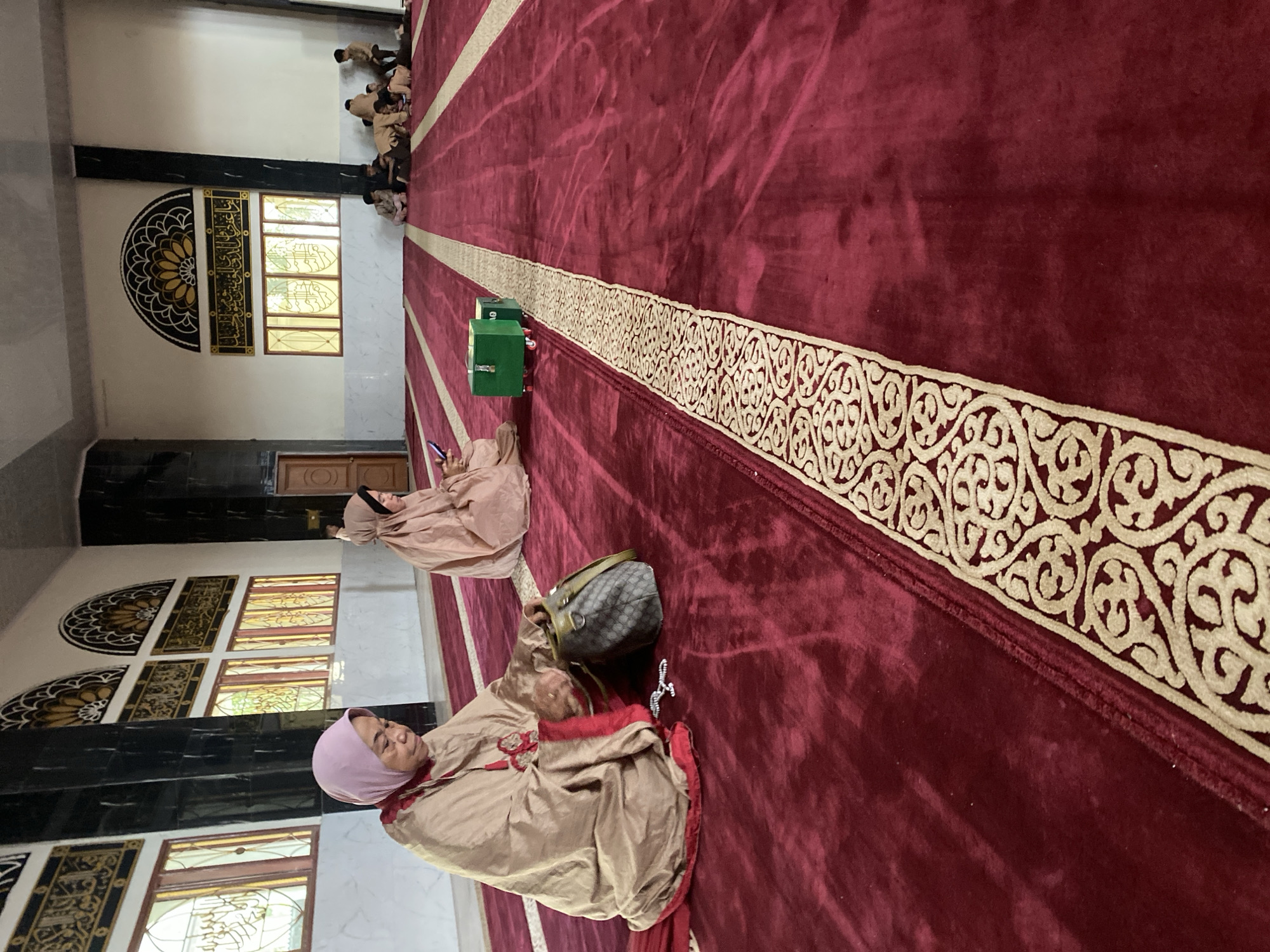 The image depicts the interior of a mosque in Manado, Indonesia, characterized by its red carpet with a decorative border and tall windows with intricate latticework. Several individuals are present, mostly women, engaged in prayer or quiet contemplation. In the foreground, a woman wearing a pink hijab and a tan robe is seen with her head bowed, hands rummaging through a dark-colored purse. Beside her lies a string of prayer beads. Further into the mosque, another woman in a similar tan garment is looking at her phone. In the background, a group of young boys are seated on the floor, seemingly engaged in conversation or activity. Two green boxes with wheels are positioned on the carpet. The mosque's architecture includes large pillars and arched windows adorned with Islamic calligraphy. The overall atmosphere is serene and reflective.