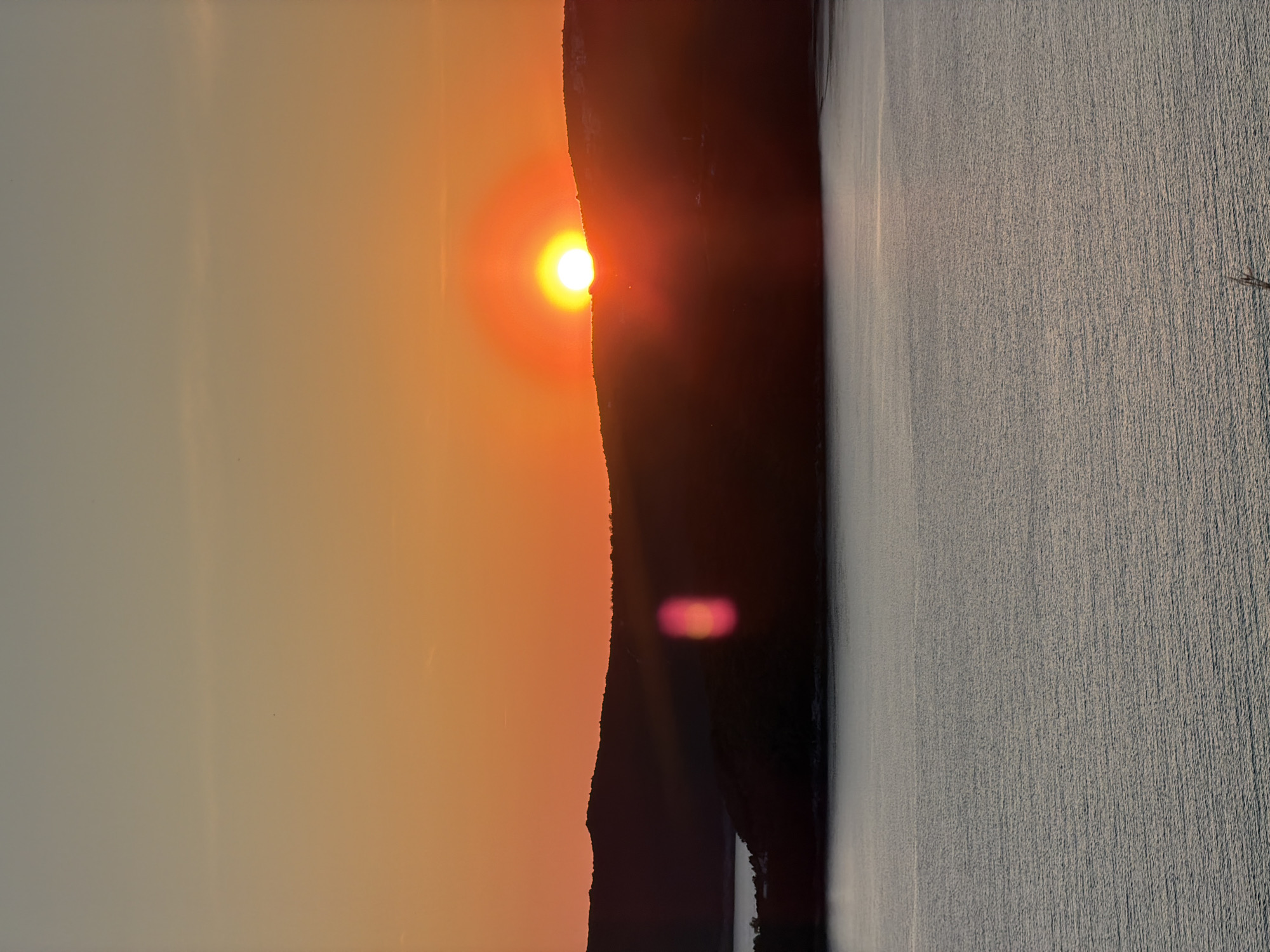The image captures a sunset in Budva, Montenegro. The sun is partially obscured by a dark, silhouetted mountain range. To the right of the mountains appears to be a body of water. The sky is a gradient of orange and gray.