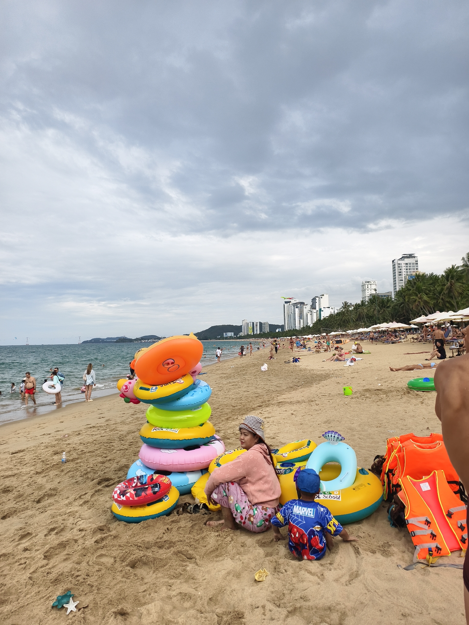 This image depicts a bustling beach scene under a heavily overcast sky. In the foreground, a woman in a pink top and patterned pants sits on the sand next to a young child wearing a blue shirt with "MARVEL" visible on it, and a blue baseball cap. They are situated amidst a collection of colorful beach accessories, prominently featuring a tall stack of inflatable swimming rings in orange, yellow, blue, pink, and green. More inflatable rings, including one marked "POOL School", are scattered around them, along with a small red and black inflatable boat. To the right, a stack of bright orange life vests is also visible.
Further along the sandy beach, numerous people are enjoying the day. Some individuals are wading or swimming in the ocean, while others walk along the shoreline. In the mid-ground, many beachgoers are relaxing on lounge chairs under white umbrellas, suggesting a resort or public beach area with amenities.
The background reveals a modern city skyline with several tall buildings and lush green palm trees lining the coast. Distant hills or mountains are visible across the water, which blends with the grey, cloudy horizon. The heavy cloud cover suggests a cool or overcast day, rather than bright sunshine. The overall atmosphere is one of leisure and recreation at a popular coastal destination.