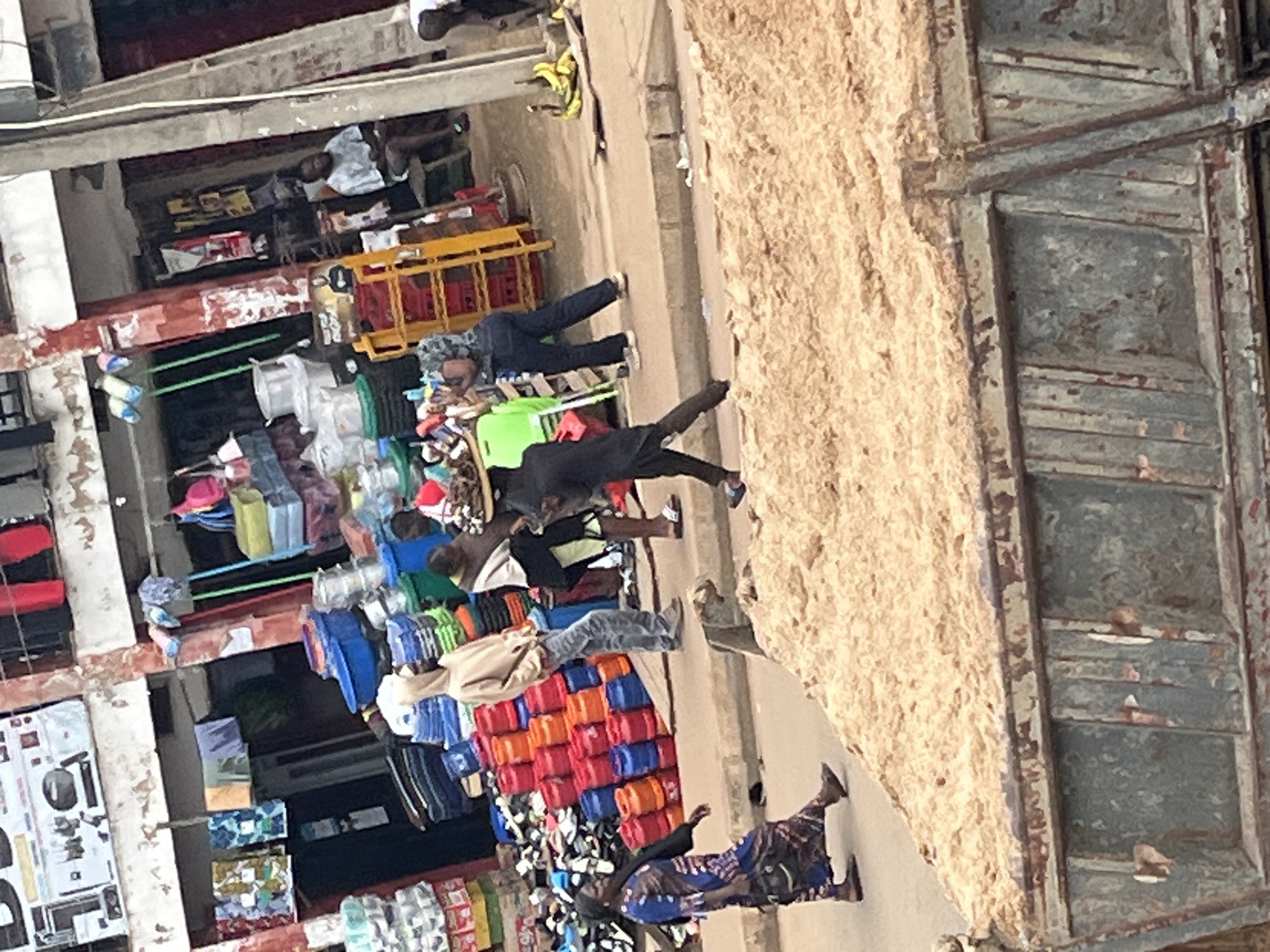 The image depicts a bustling street scene in Bukuru, Nigeria, likely a market area. In the foreground, a large, weathered truck bed is filled with sand, obscuring the lower portion of the scene. Behind the truck, a group of people are engaged in various activities.
A woman in a blue and brown patterned dress walks from left to right, her head covered by a black scarf, carrying a black bag on her shoulder. To her right, a man wearing a light brown hooded sweatshirt and grey jeans stands looking towards the right. Further to the right, a person in a black outfit is carrying a tray of goods on their head. They are walking from right to left.
In the background, market stalls are visible, with shelves stocked with various items, including plastic basins, stacks of colorful containers (red, orange, and blue), and what appear to be fabrics or clothing hanging from a line. Some individuals are seen sitting or standing near the stalls. A man in a light-colored t-shirt is seated on a stool to the right of a yellow shelf unit. A cluster of bananas is visible on the ground to the far right. The environment appears to be an outdoor market with a mix of goods for sale and pedestrian activity. The buildings in the background show signs of age, with faded paint and exposed elements.