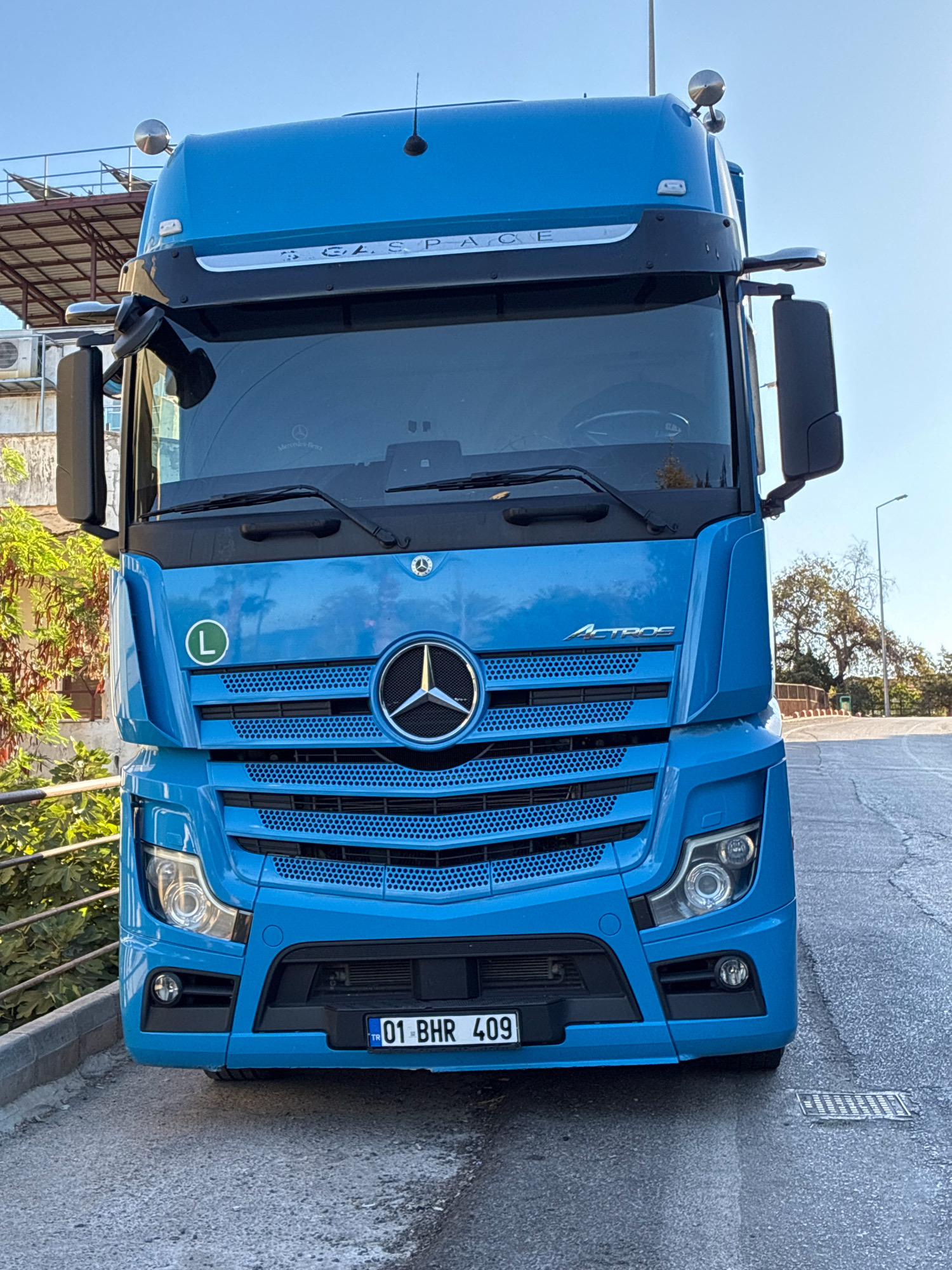 A bright blue Mercedes-Benz Actros truck is parked facing the camera on a slightly uphill asphalt road, casting a shadow. The truck's front is prominently displayed, featuring its large grille with the Mercedes-Benz star emblem, distinctive headlights, and fog lights. A license plate with the text "01 BHR 409" is visible. On the left side of the truck's front, a green circular sticker with a white "L" is attached. Above the windshield, the text "BIGSPACE" is visible. The background shows a clear blue sky, some greenery including trees and bushes to the left of the truck, and a building structure with solar panels in the upper left. The overall setting appears to be an outdoor area, possibly a roadside or parking lot, with a hint of urban elements. The location is identified as Alanya, Turkey.