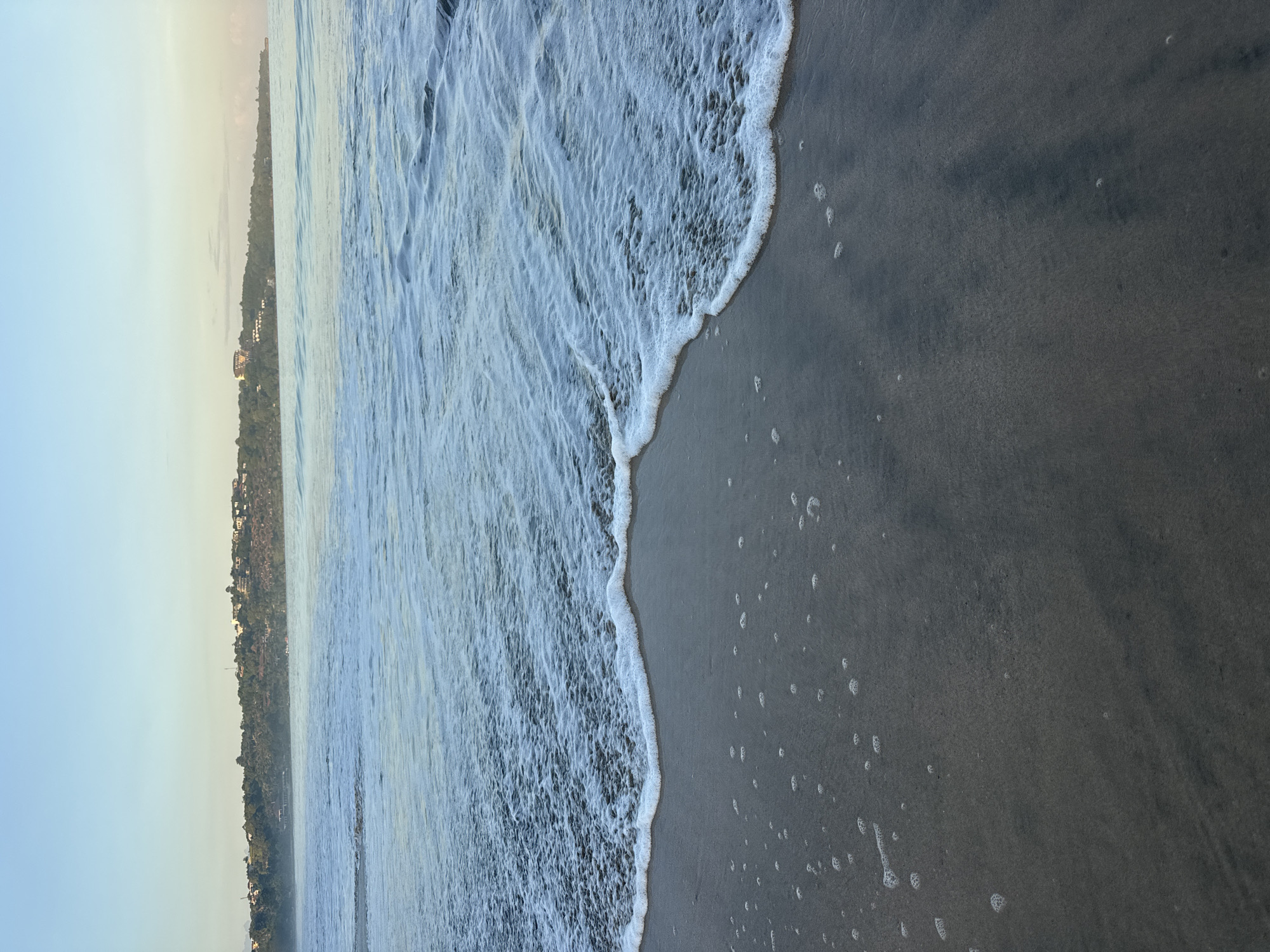 The image depicts a shoreline in Unknown, Indonesia. Gentle waves with foamy white crests are washing onto a dark grey sandy beach. In the distance, a coastline with buildings and vegetation is visible under a pale sky. The scene suggests either early morning or late evening due to the soft lighting.