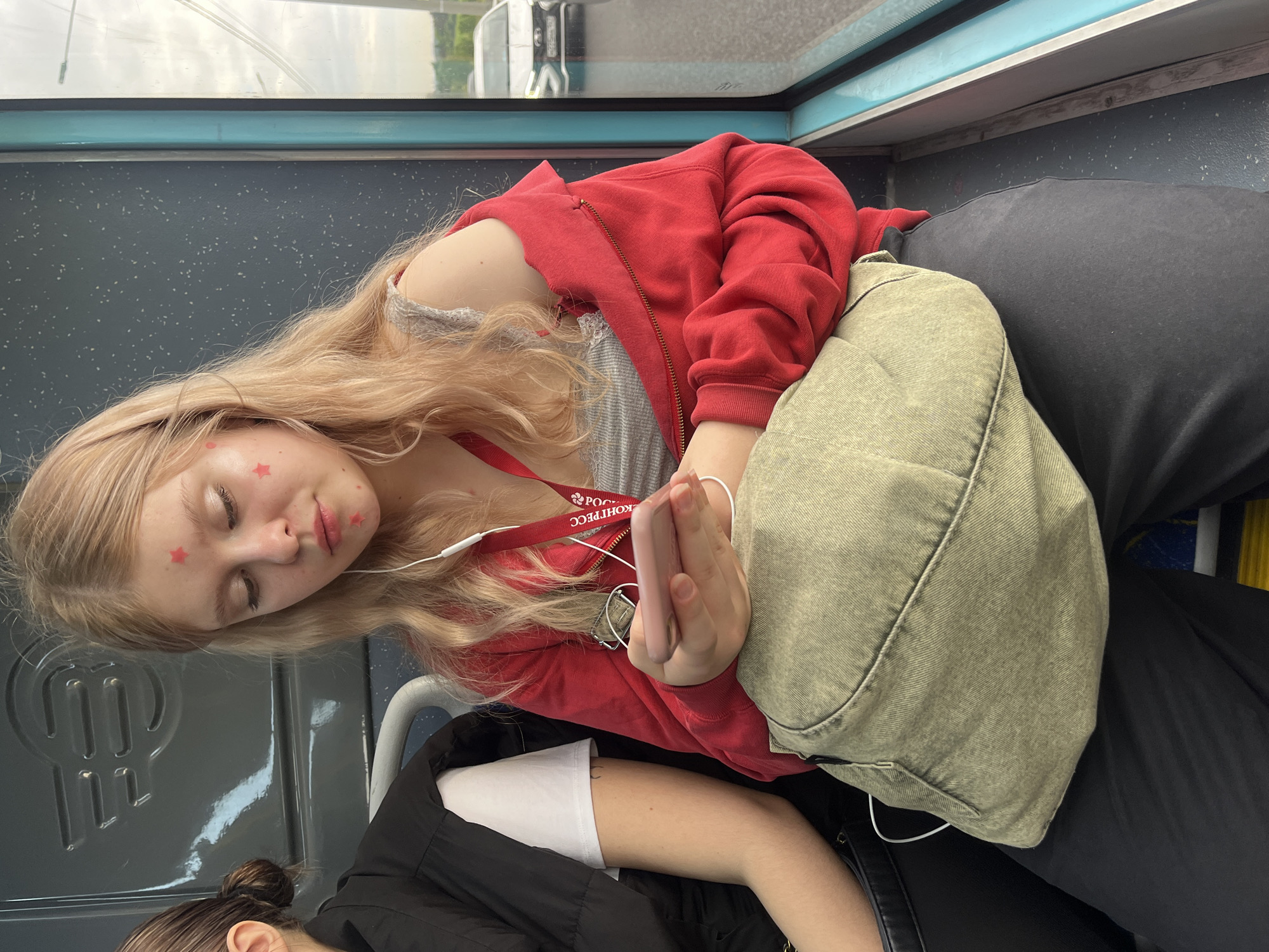 Two individuals are depicted inside what appears to be a moving vehicle, possibly a bus or train, with an overcast or rainy exterior visible through the window. The primary subject is a young person with long blonde hair, their eyes closed in repose, suggesting they are sleeping or resting. They have several small, red star-shaped stickers on their face, near their eyes and cheek. They are wearing a red hooded zipper jacket over a grey, lace-trimmed top, and dark trousers. A light olive-green fabric shoulder bag rests on their lap, and they hold a pink smartphone with clasped hands. White wired earbuds are visible, connected to the phone. A red lanyard, bearing the Cyrillic text "РОСКОНГРЕСС" (ROSKONGRESS), hangs around their neck.
Partially visible in the lower part of the frame is another person, seen from behind, with dark hair tied in a bun. This individual is wearing a black jacket over a white top and appears to be resting or sitting.
The interior of the vehicle features dark, speckled walls and a light blue trim along the window line. A grey plastic panel with an embossed design is visible near the second person's head. The overall scene conveys a quiet moment of travel or commuting.
