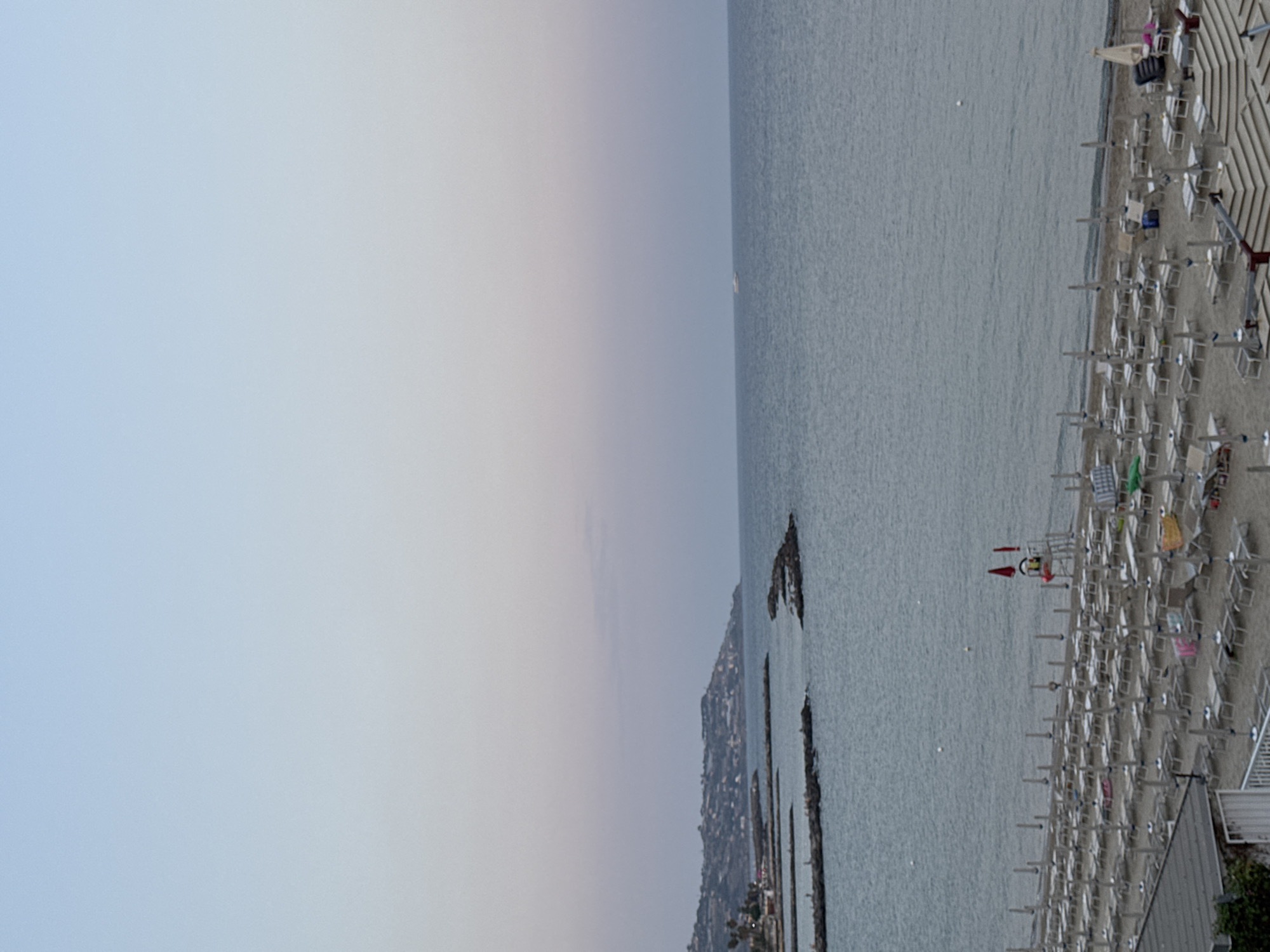 The image depicts a beach in Sanremo, Italy, during twilight. The sky is a soft gradient of blue and pink. The calm sea stretches to the horizon, where a small white boat is visible. Along the coastline, a hilly landscape dotted with buildings is present. In the foreground, rows of empty lounge chairs and umbrellas are neatly arranged on the sandy beach. A lifeguard station with red flags is situated on the sand. The scene conveys a peaceful and serene atmosphere.