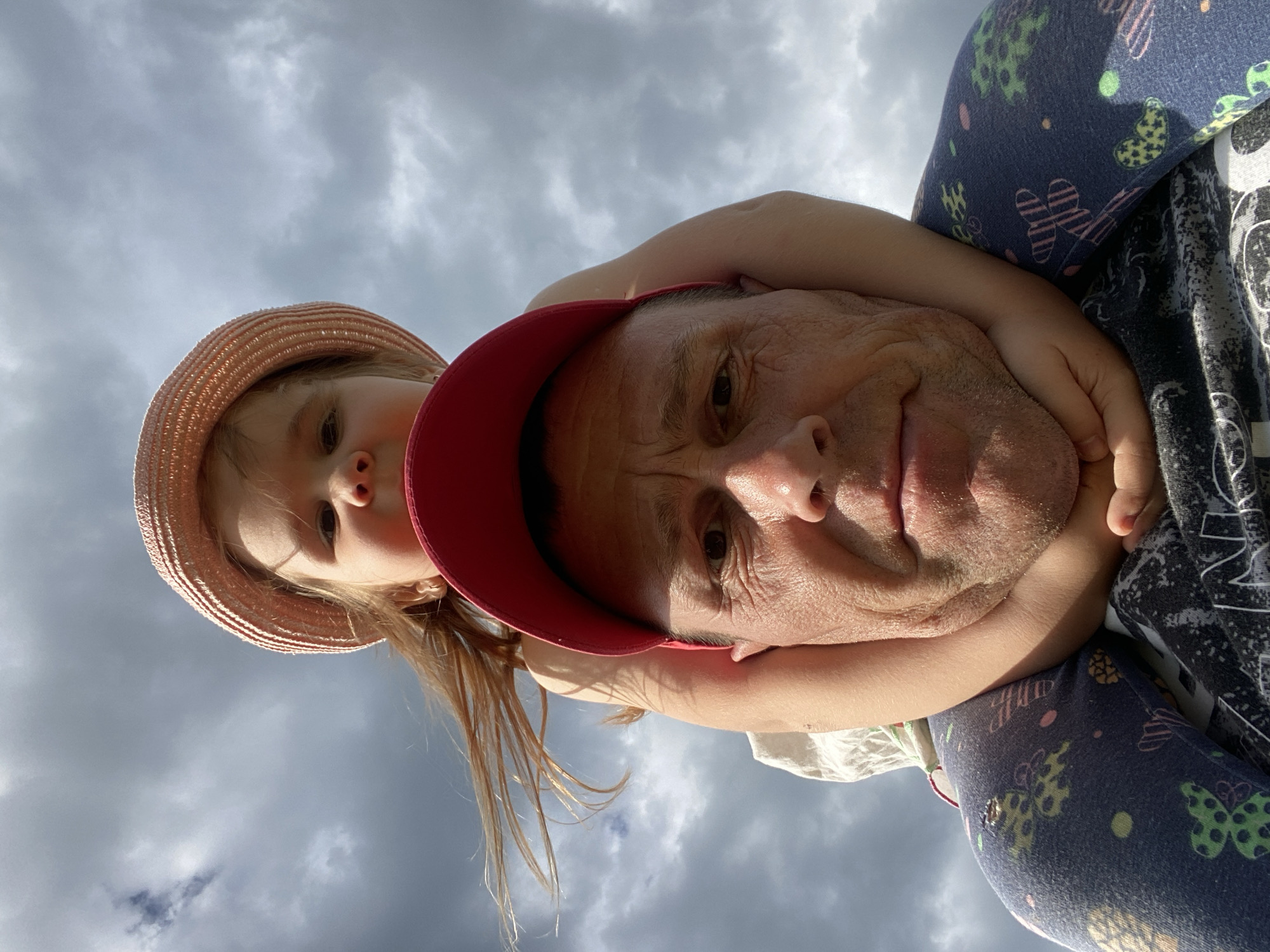 A low-angle, upward-facing photograph shows a man and a young girl in an embrace against a cloudy sky. The man wears a red baseball cap and is seen from the neck up. The girl, wearing a straw hat, is behind him, her arms wrapped around his neck. The location cannot be determined from the image.