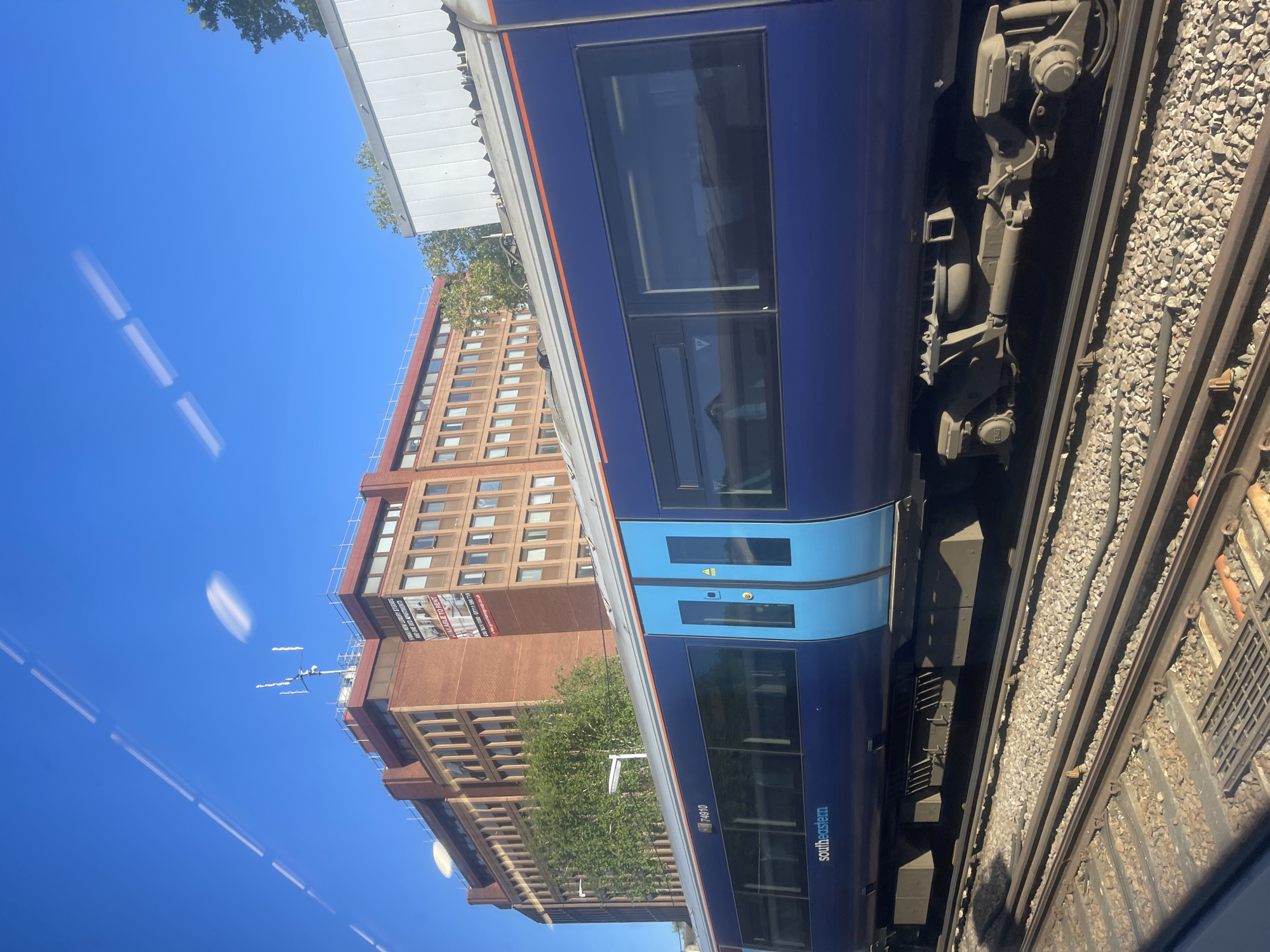 The image, originally oriented 90 degrees clockwise, captures a bright, sunny daytime scene from the perspective of a train window. The clear blue sky dominates the upper and left portions of the frame, showing faint reflections of interior lighting on the glass.
On the right side, a dark blue Southeastern train is prominently featured, identifiable by its "southeastern" branding and unit number "74910". It has contrasting light blue doors. Below the train, detailed railway tracks with gravel ballast are visible, along with some of the train's mechanical undercarriage components.
In the background, a tall, multi-story building constructed of reddish-brown brick stands against the sky on the left. It features many windows and what appears to be scaffolding or an antenna array near its roofline. A large banner on the building advertises "BRAND NEW LUXURY STUDIOS AND ONE BED APARTMENTS AVAILABLE TO RENT" and provides contact numbers: "020 3230 0080" and "07711 590 719". Green foliage from trees is visible between the train and the building. The scene conveys a clear, bright day in Tonbridge and Malling, United Kingdom.