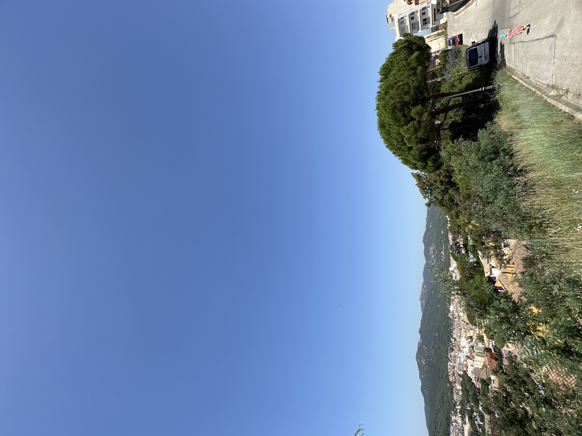 The image captures a sun-drenched day in Sant Feliu de Guíxols, Spain, with a clear, expansive blue sky dominating the upper portion of the frame. In the foreground, a paved pathway slopes downwards, bordered by tall, dry grasses and a large, dense pine tree with a rounded canopy. To the right of the pathway, a white building with multiple balconies is visible. On the pathway itself, a silver van is parked, and further down, a person wearing a red dress and a striped scarf is walking away from the viewer. Two cars, one red and one dark-colored, are parked on the street beyond the pathway.
In the middle ground, a panoramic view unfolds of the coastal town of Sant Feliu de Guíxols. Buildings with terracotta roofs and white facades are clustered together, nestled against a backdrop of lush green hills covered in trees. The distant mountains form a rugged horizon against the clear sky. The overall impression is one of a bright, summer day in a Mediterranean coastal town.