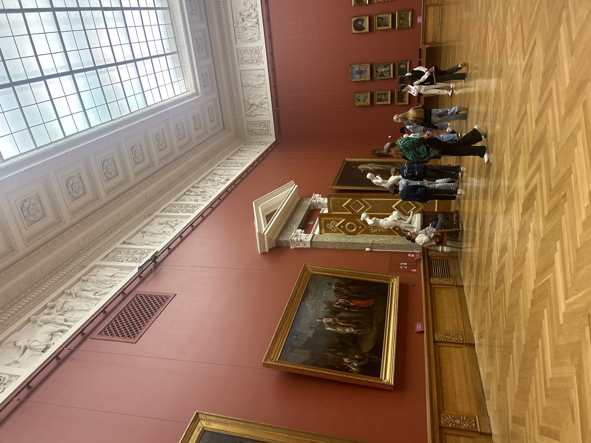 This is an interior view of the State Hermitage Museum in Saint Petersburg, Russia. The image showcases a grand art gallery with high, ornate ceilings featuring a skylight that illuminates the space. The walls are painted a deep red, adorned with numerous framed paintings and classical sculptures. In the foreground, a group of visitors, dressed casually, are walking and observing the artwork. One visitor is seated on a chair near a large painting. The floor is made of polished light-colored wood laid in a herringbone pattern. The overall scene depicts a typical museum environment with people engaging with art.